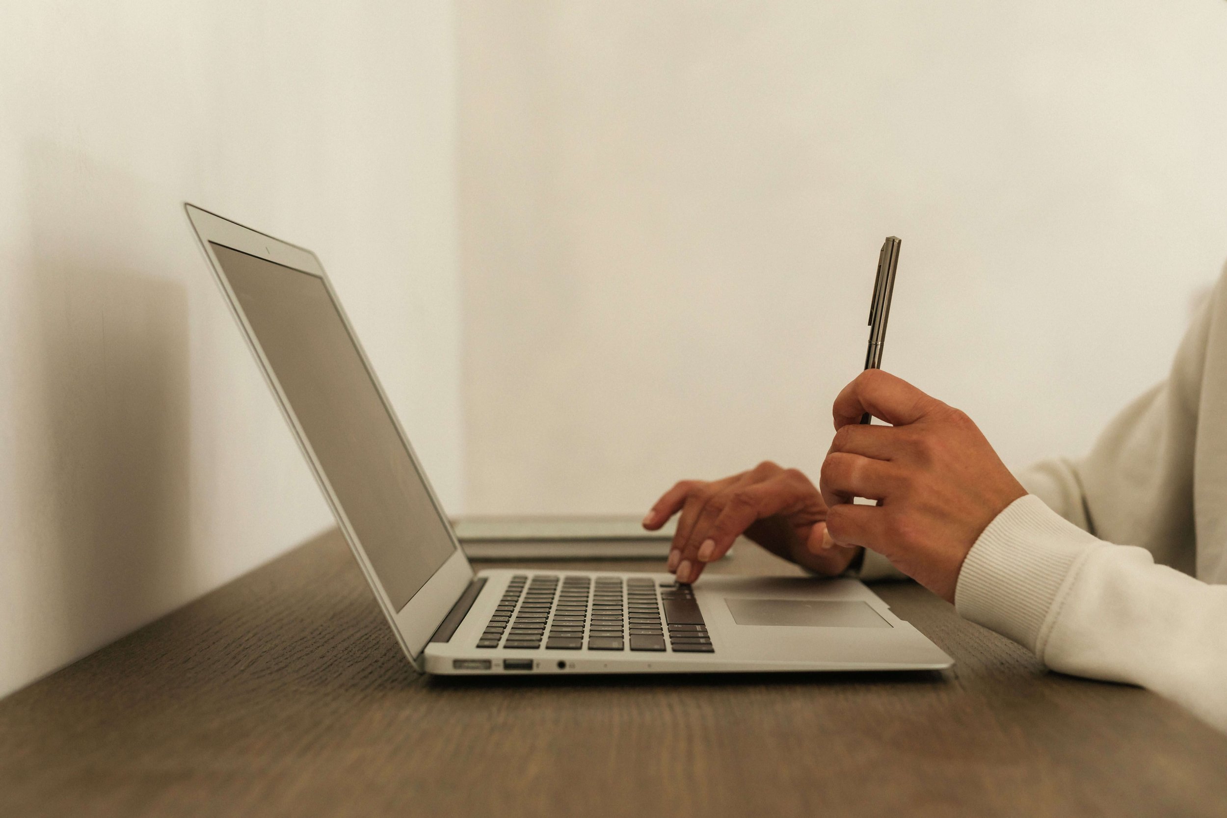 Person using a laptop and holding a pen at a wooden desk against a plain white wall.