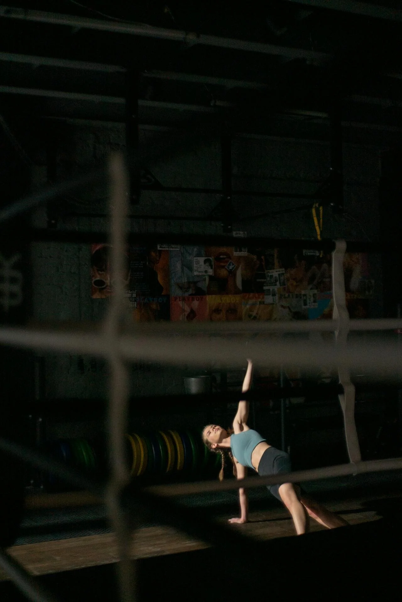 A woman in a gym performing a downward dog yoga pose, viewed through a metal rack in a dimly lit space with workout equipment.