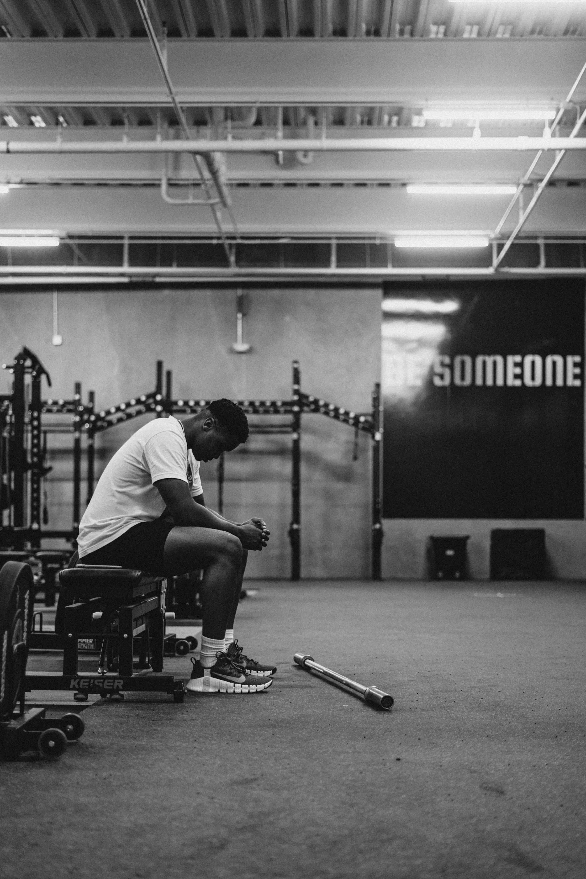 A person sitting on a bench inside a gym, looking down at their hands. The gym has workout equipment and a sign on the wall that partially reads 'SOMEONE'.