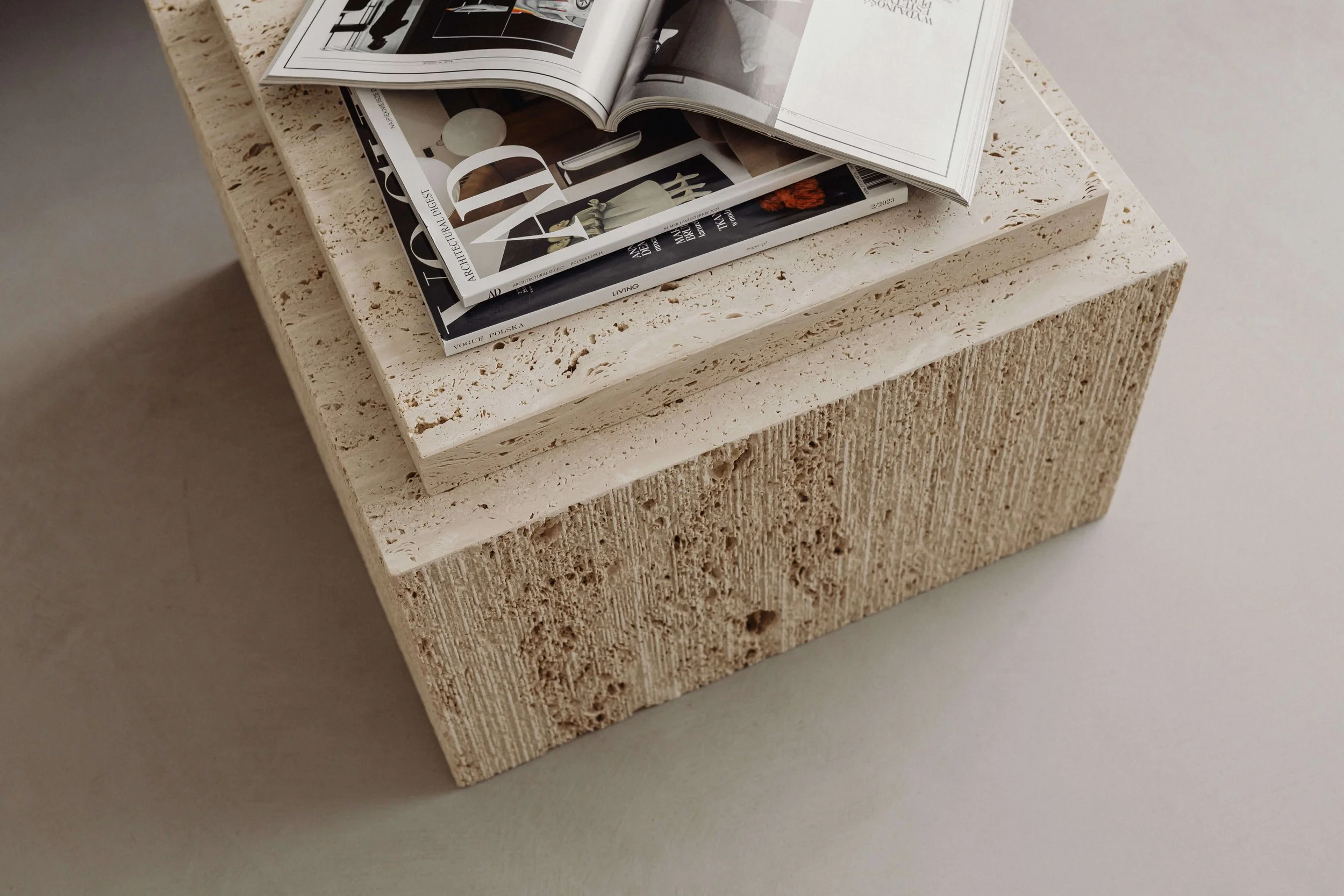 A close-up of a beige textured block with a flat top, stacked with modern magazine and book pages, featuring architectural and home decor images, on a light surface.