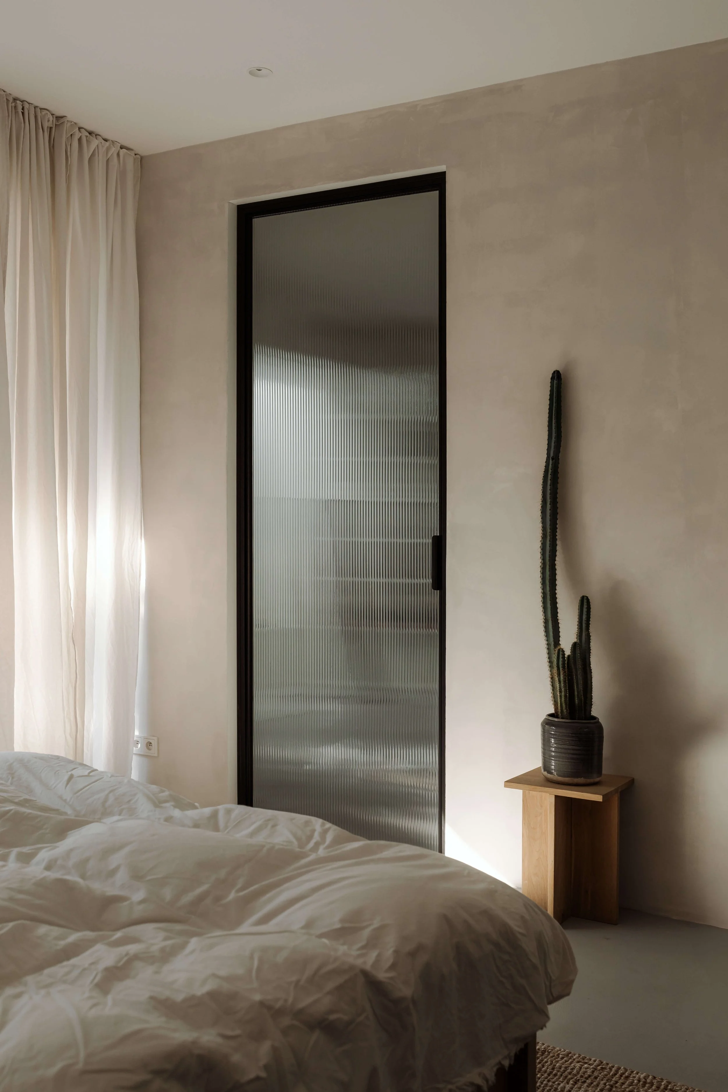 A bedroom with beige walls, a bed with white bedding, a tall potted cactus on a small wooden side table, and a frosted glass sliding door, with cream curtains on the window.