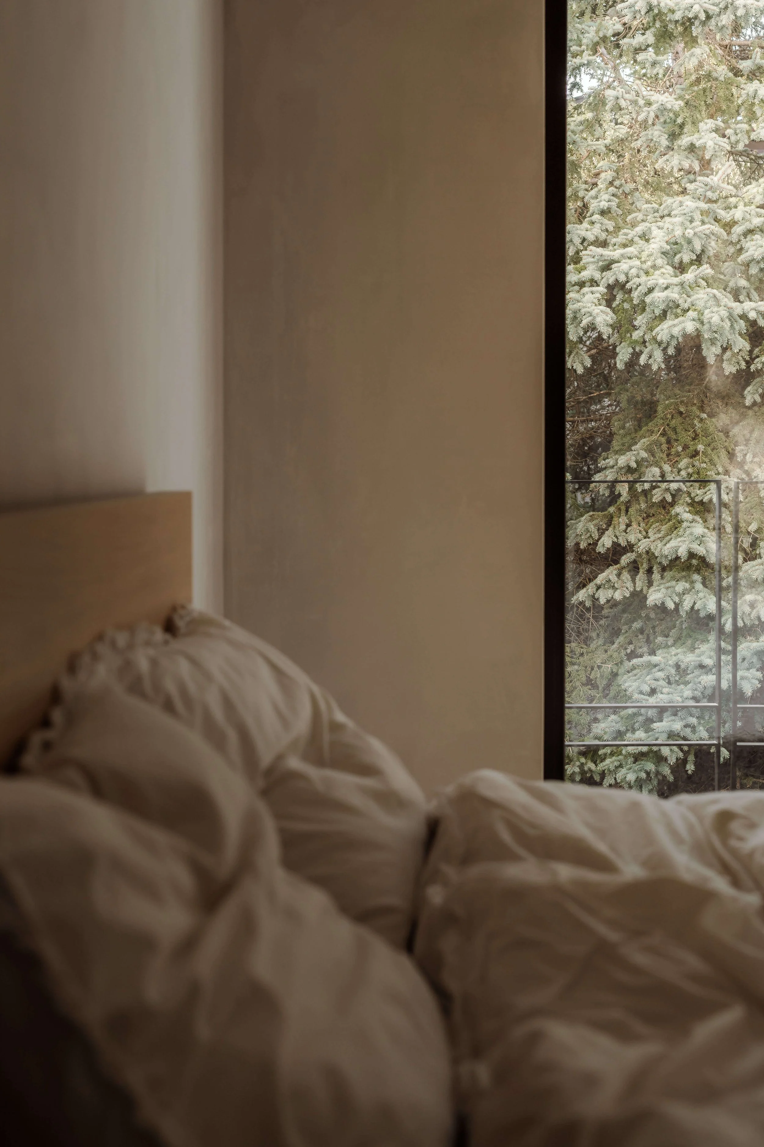 Unmade bed with white rumpled sheets and pillows near a window showing snow-covered evergreen trees outside.