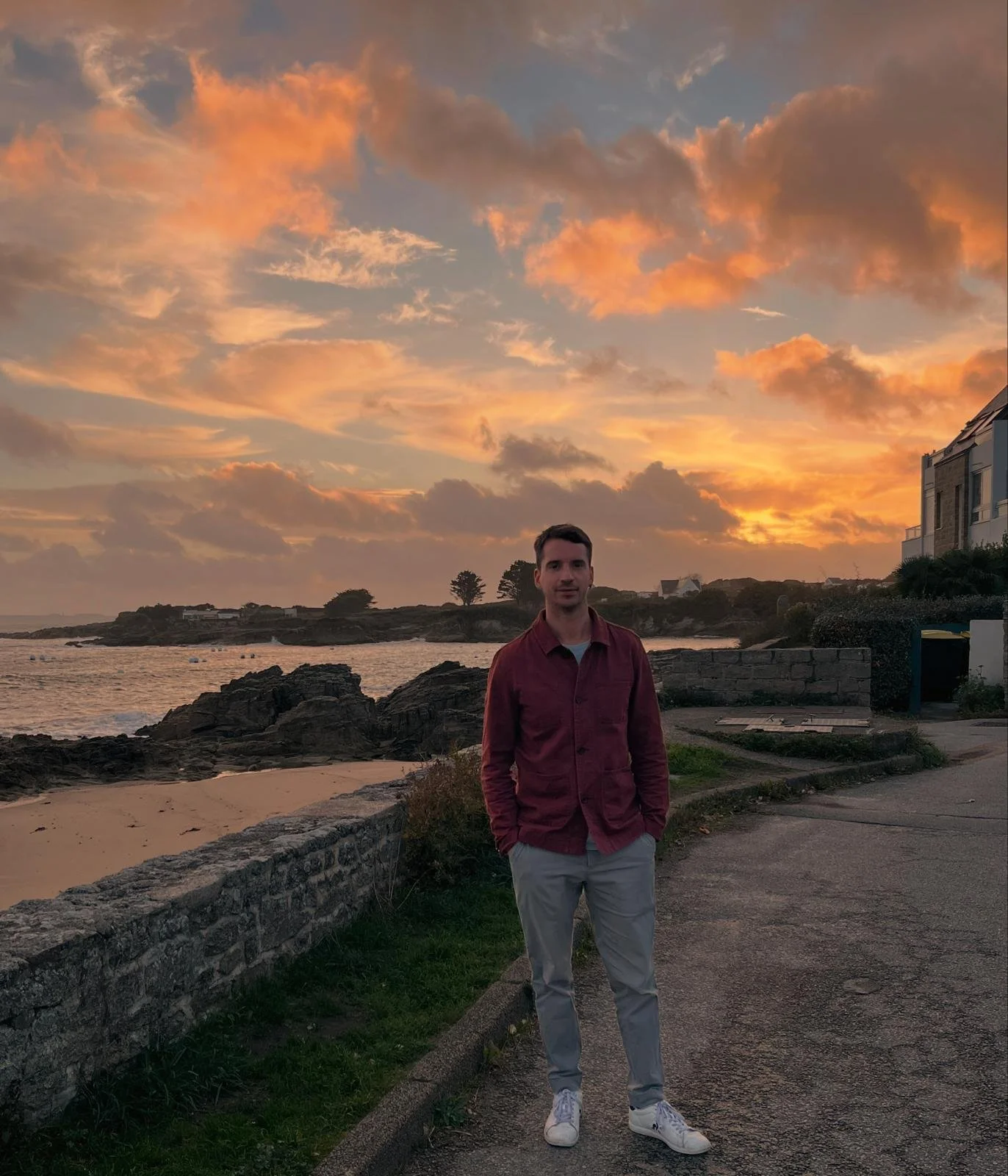A young man standing on a pathway near a rocky beach at sunset, with a partly cloudy sky and residential buildings in the background.