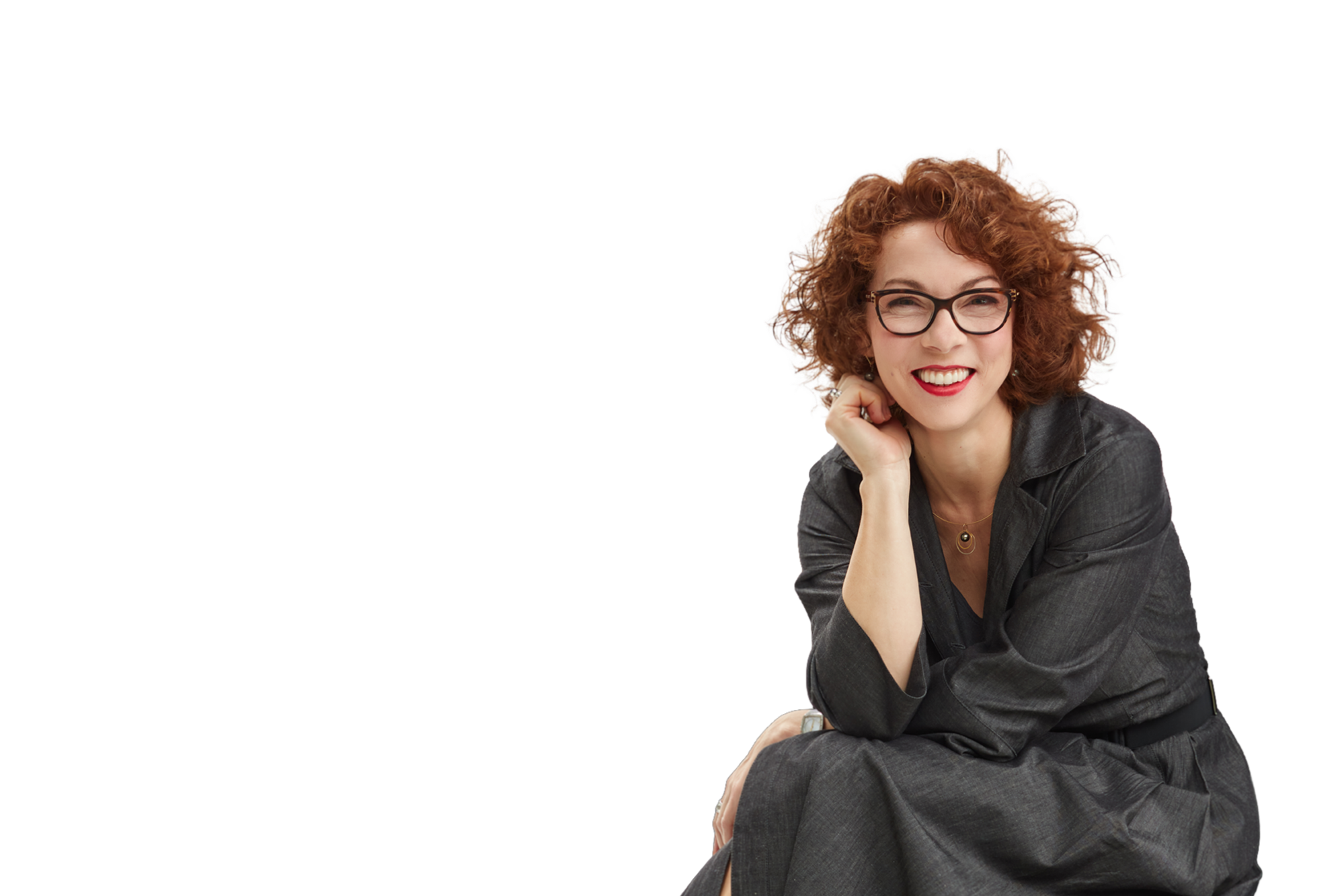 A woman with curly red hair, glasses, and red lipstick sitting with her chin resting on her hand, smiling at the camera.