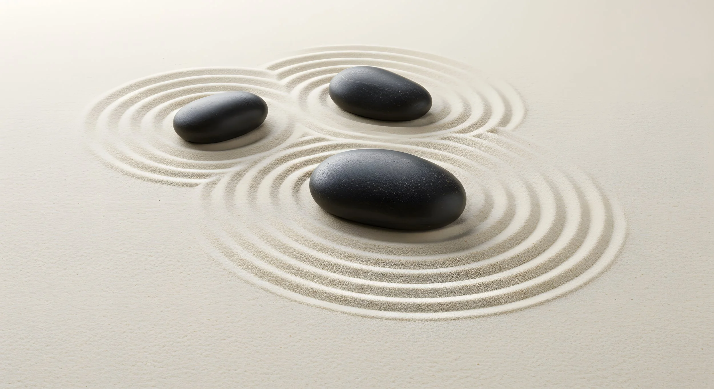Three smooth black stones placed on a white surface with concentric sand patterns around each stone, resembling a Zen or meditation setting.