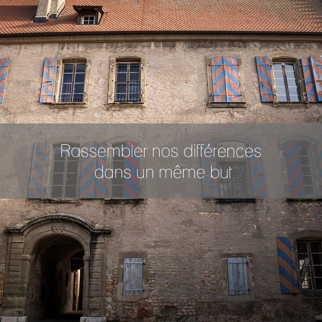 Une façade d'un vieux bâtiment en pierre avec fenêtres à volets bleus et rouges, un porte en pierre sculptée, et un toit en tuiles.