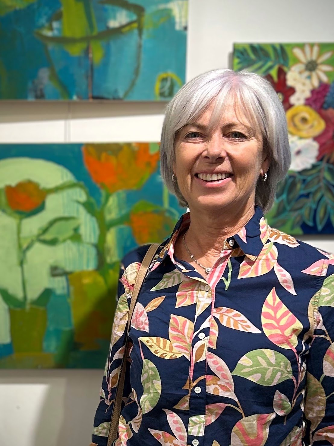 A smiling older woman with short gray hair wearing a navy blouse with colorful leaf patterns, standing in front of vibrant floral paintings at an art gallery.