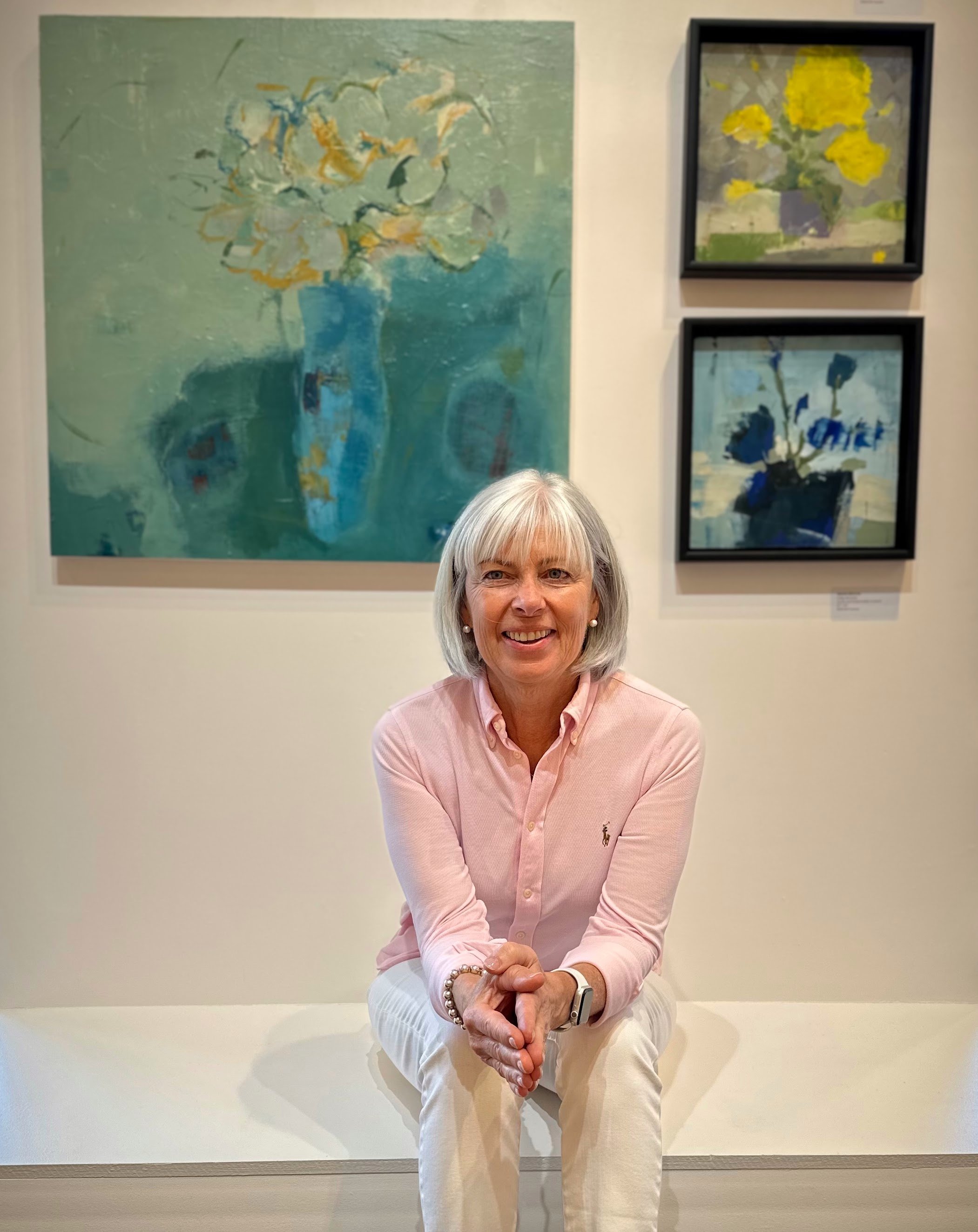 A woman with gray hair, wearing a pink button-up shirt and white pants, sitting on a bench in an art gallery, with three paintings of flower arrangements behind her.