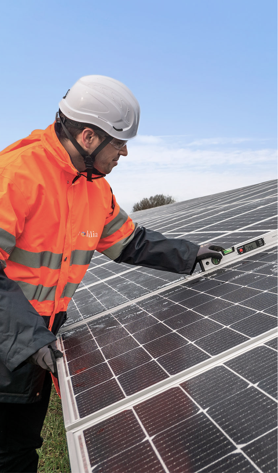 A man wearing a white safety helmet and an orange safety jacket inspects solar panels on a solar farm using a handheld device.