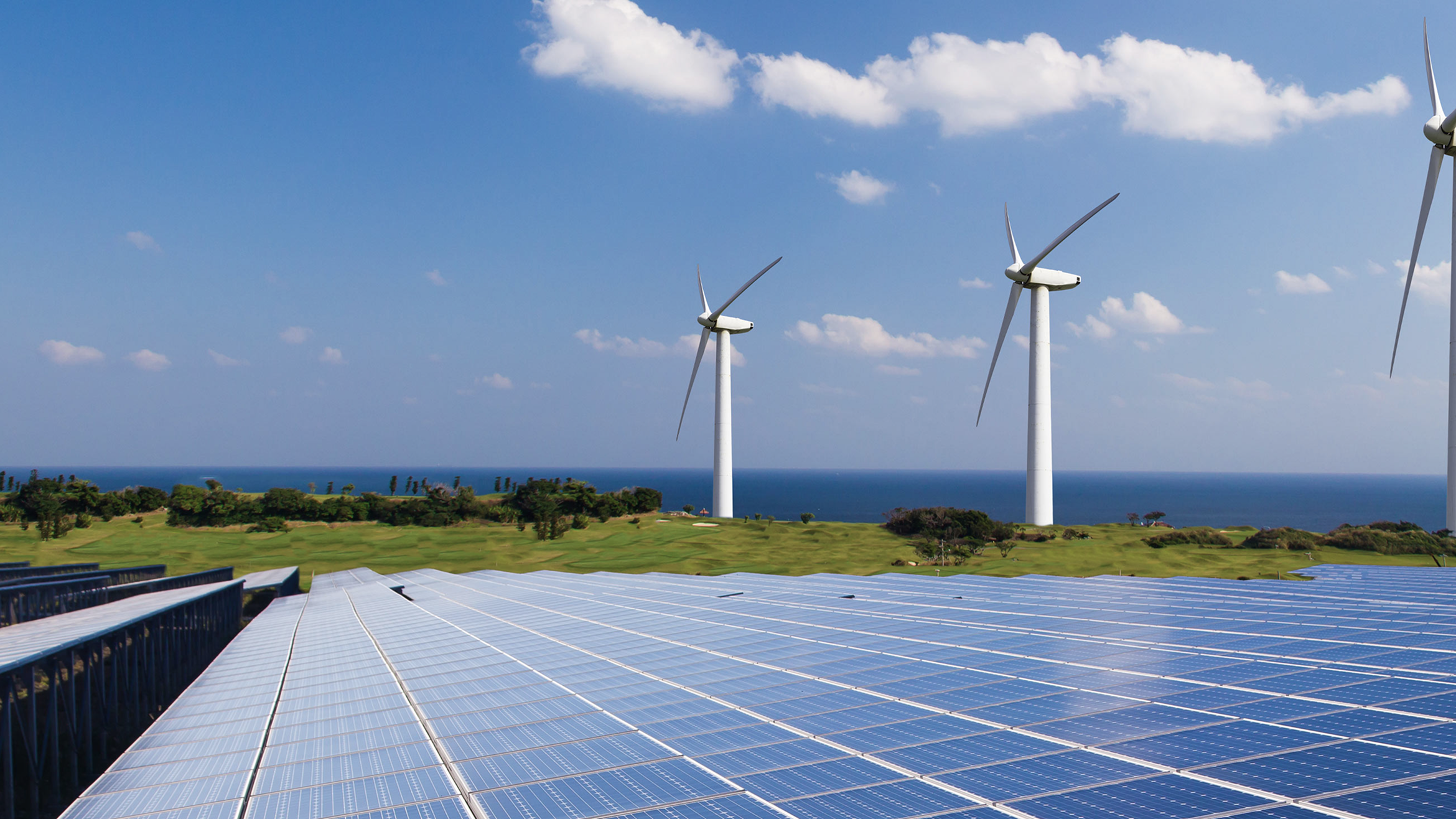 View of solar panels in the foreground with wind turbines and ocean in the background under a partly cloudy sky.