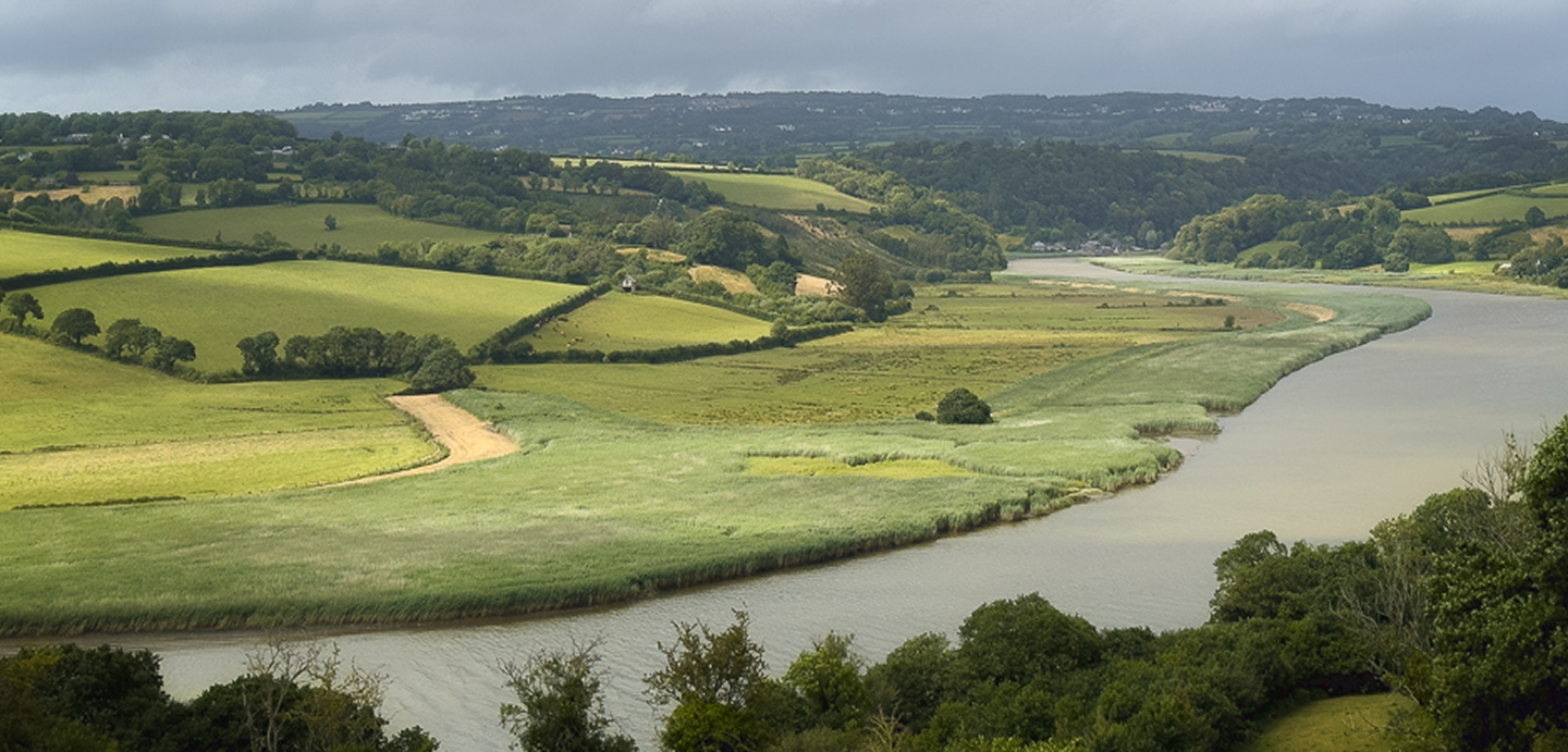 A scenic view of a winding river flowing through lush green fields and rolling hills under a cloudy sky.