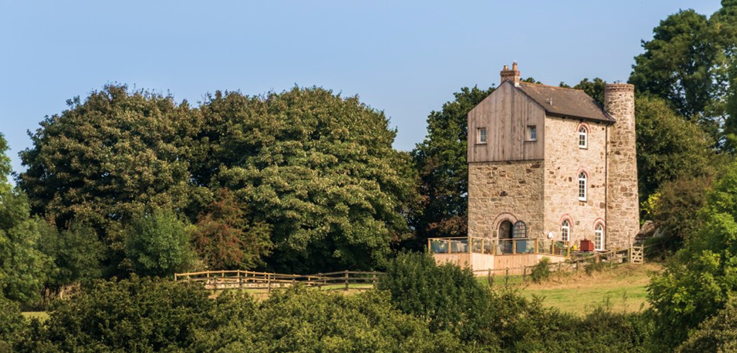 A large stone house with a wooden section on top, surrounded by lush green trees and a grassy yard, under a clear blue sky.