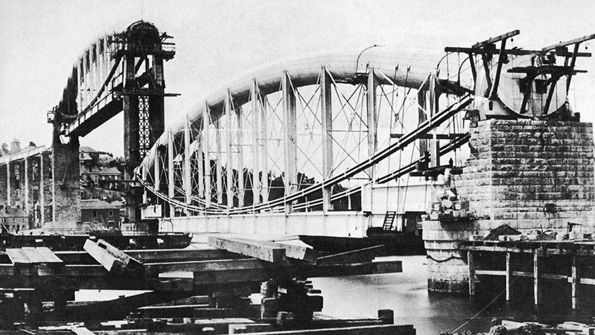 Historical black and white photograph of the construction of the Sydney Harbour Bridge, featuring the steel arch, supporting pillars, and construction equipment.