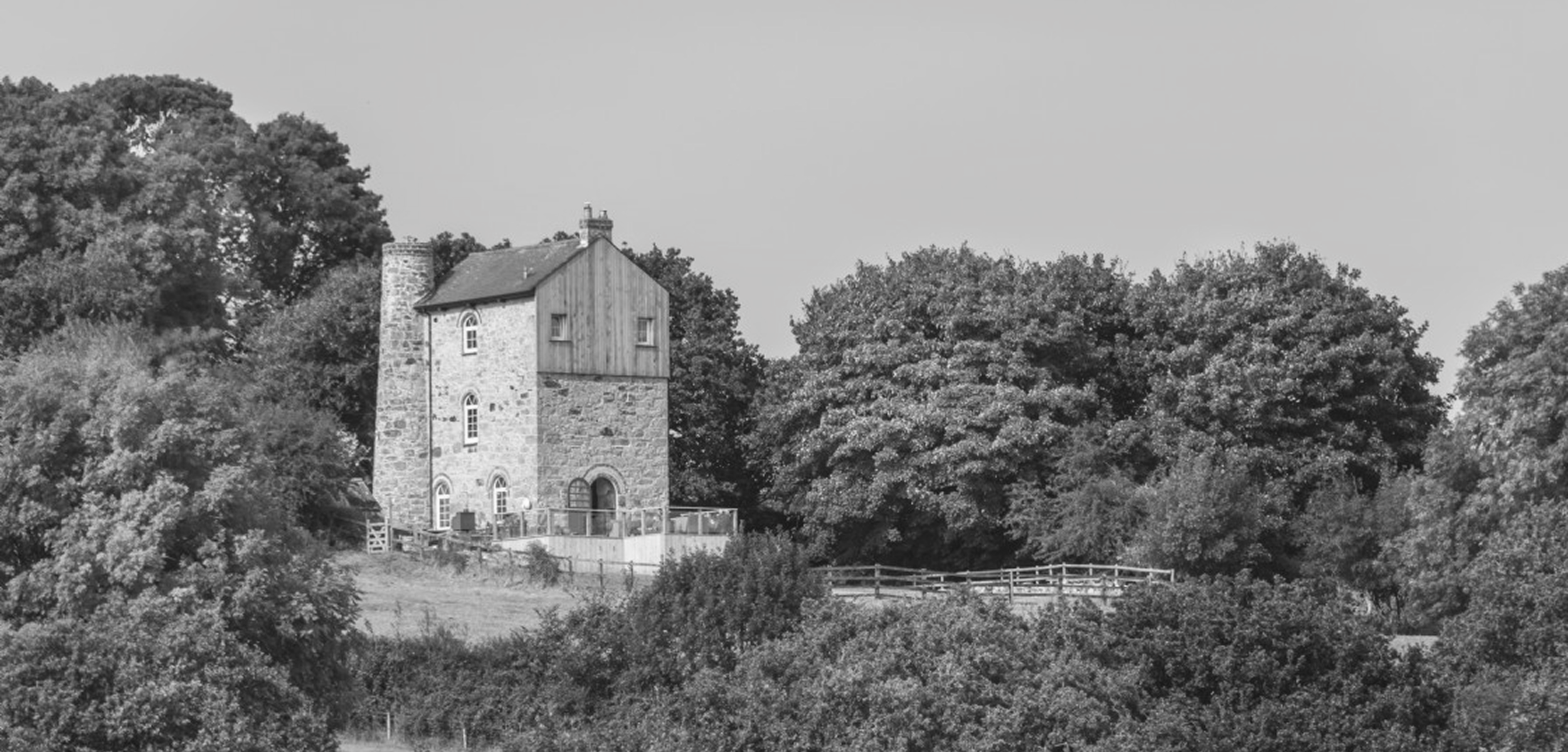 A stone house with a wooden upper level and a chimney, surrounded by trees and a wooden fence.
