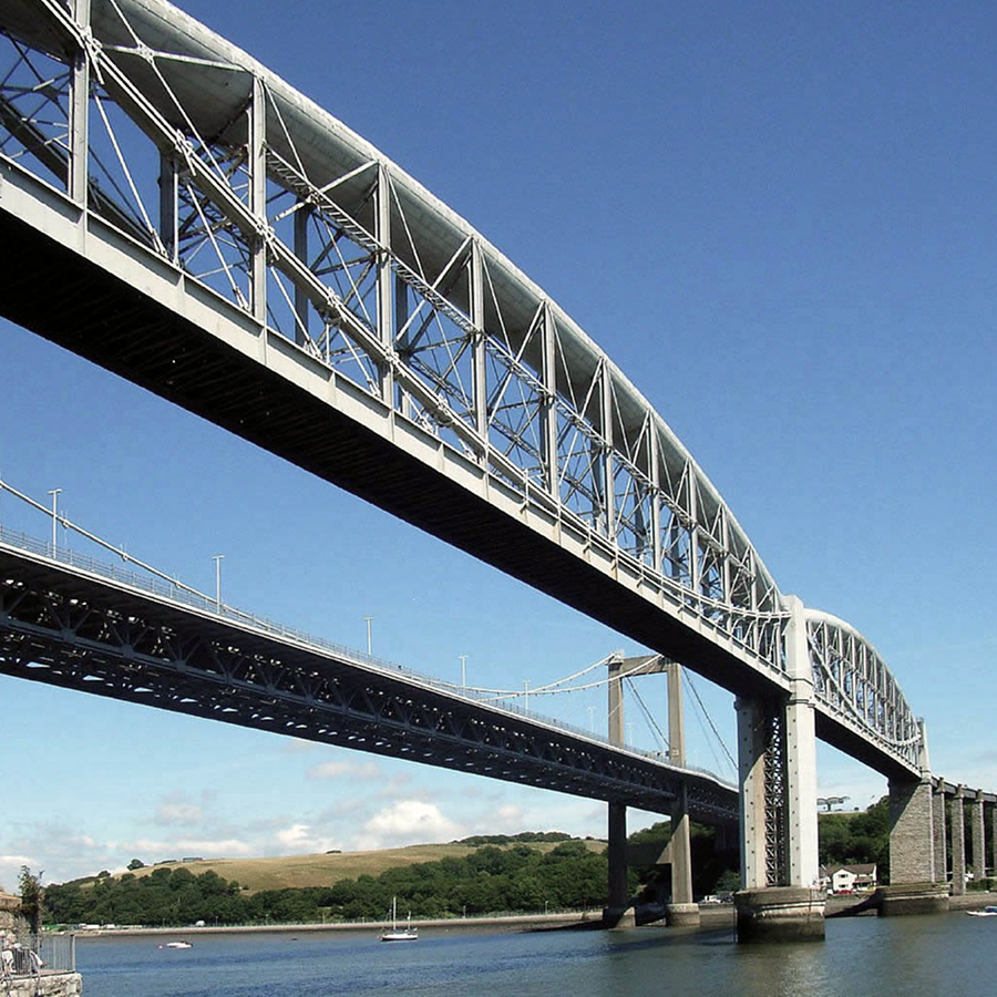 Steel bridge spanning over a body of water with a hilly landscape in the background under a clear blue sky.