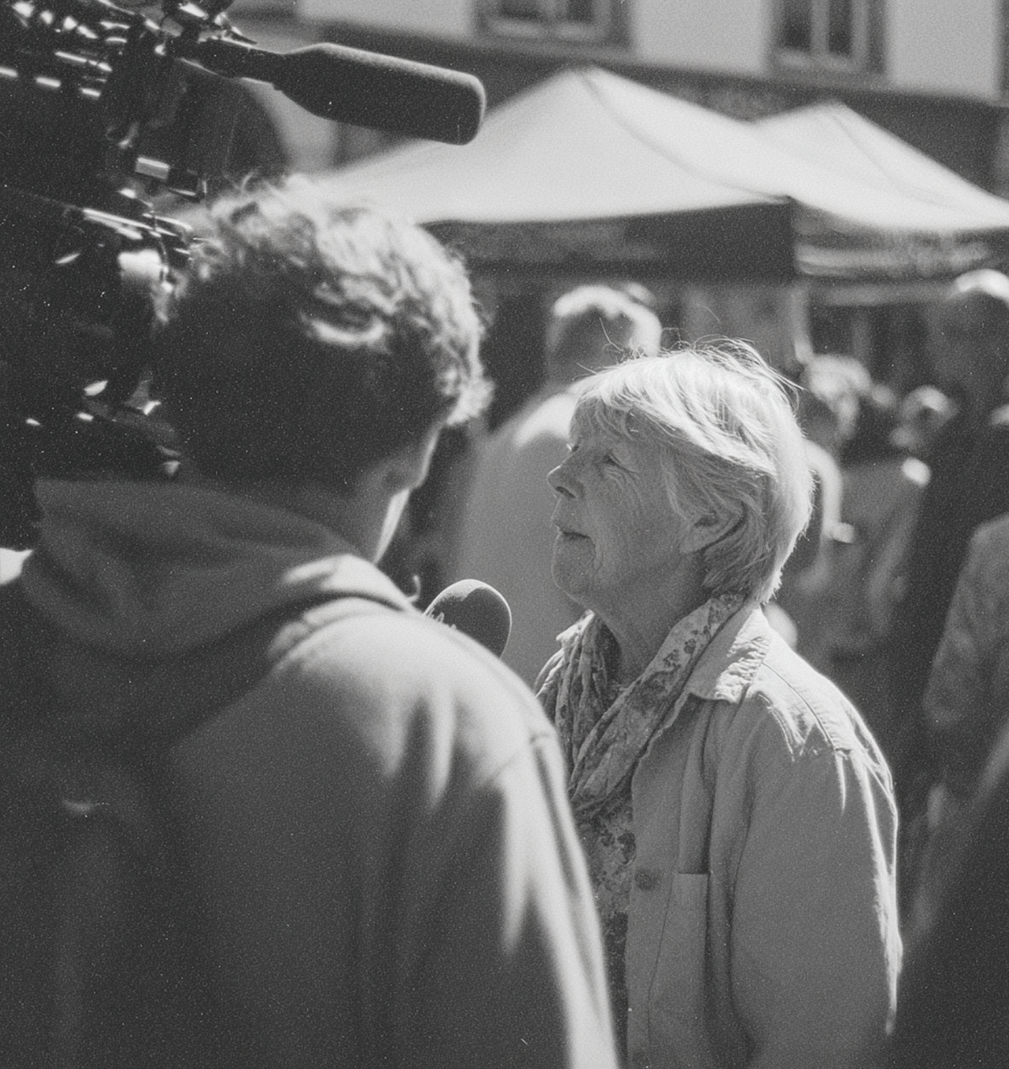 An elderly woman being interviewed by a person with a camera on a busy street with tents.
