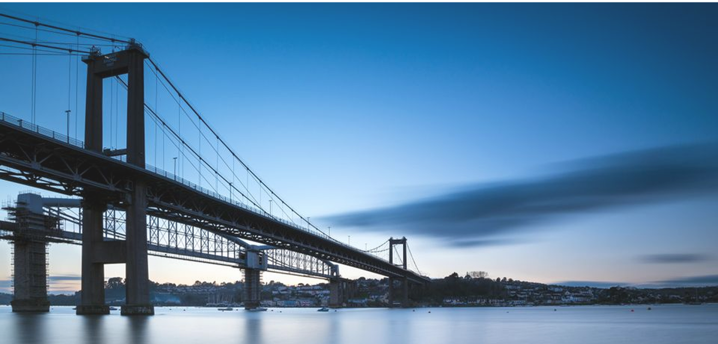 View of a large suspension bridge over water at dusk with a cityscape in the background.