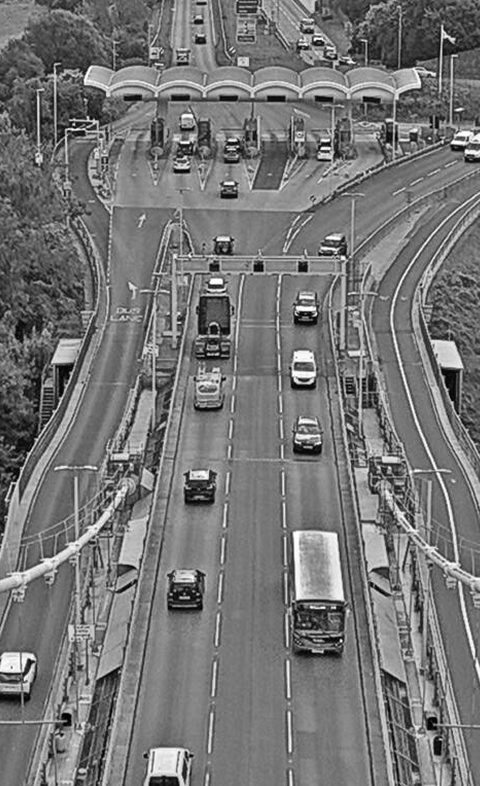 Multiple vehicles, including cars and buses, on a multi-lane highway near a toll booth with a canopy, in an urban area.