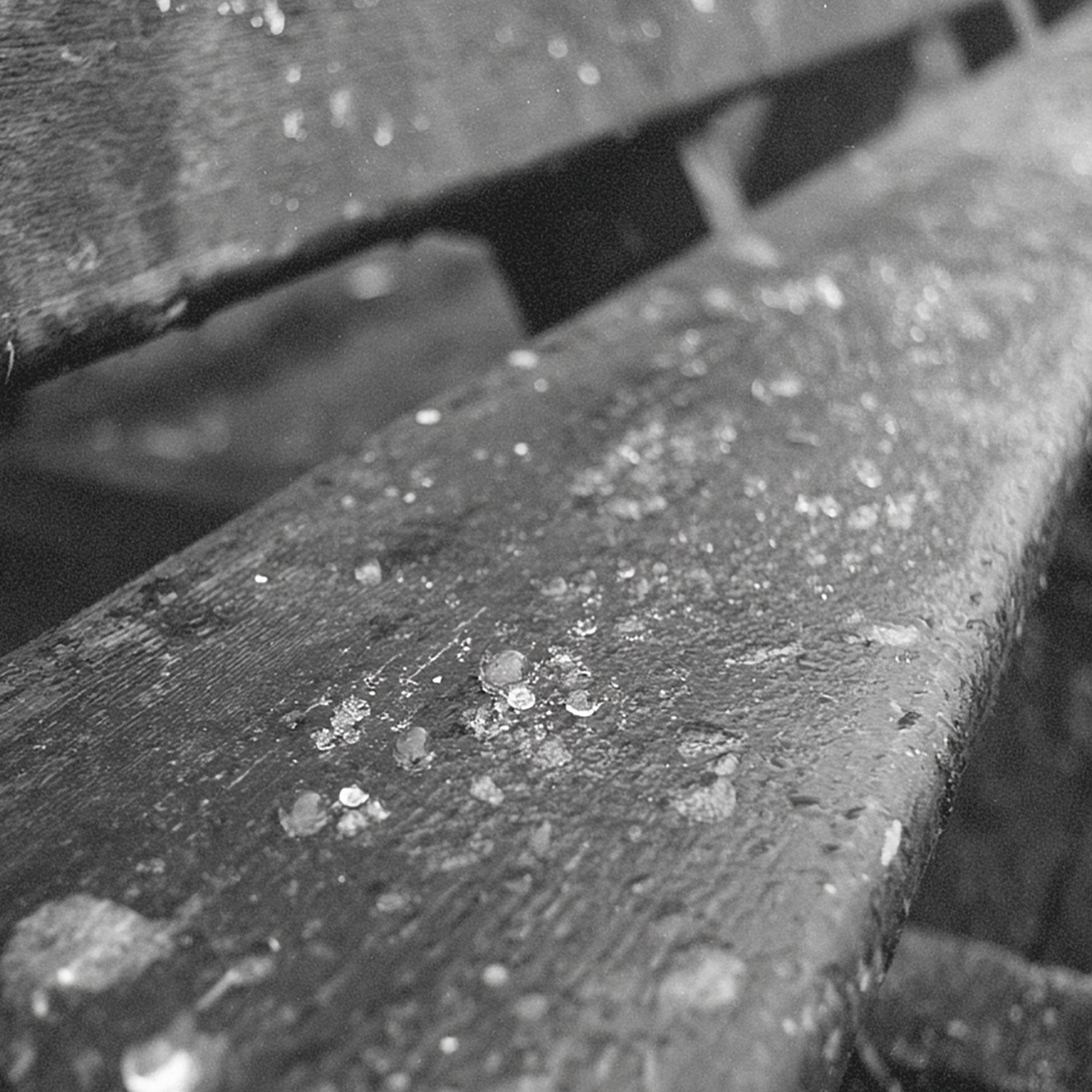 Close-up black and white photo of raindrops on a wooden surface, possibly a bench.