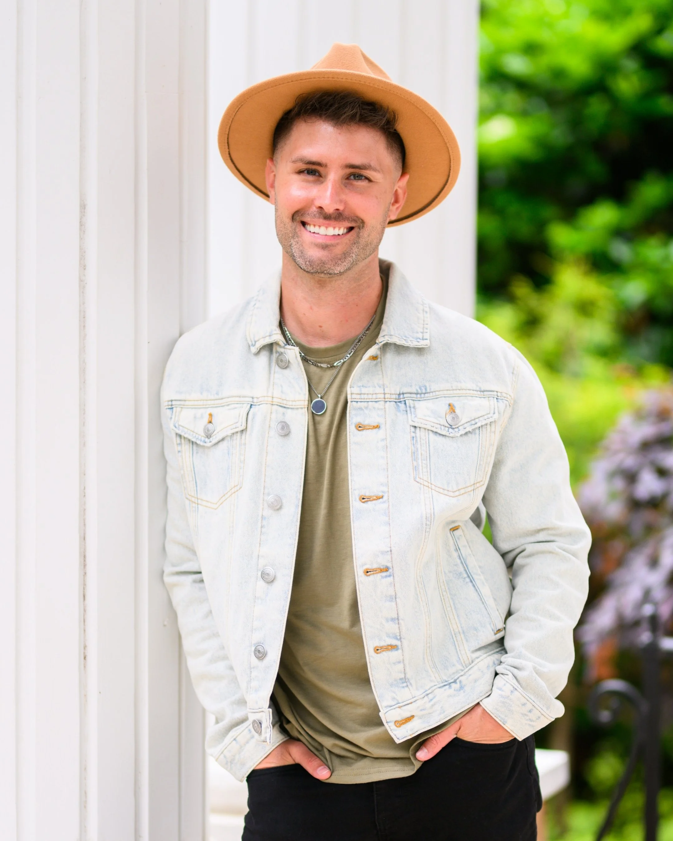 A man wearing a beige fedora hat, a light denim jacket, and a necklace with a round pendant, smiling and standing outdoors near a white wall with greenery in the background.