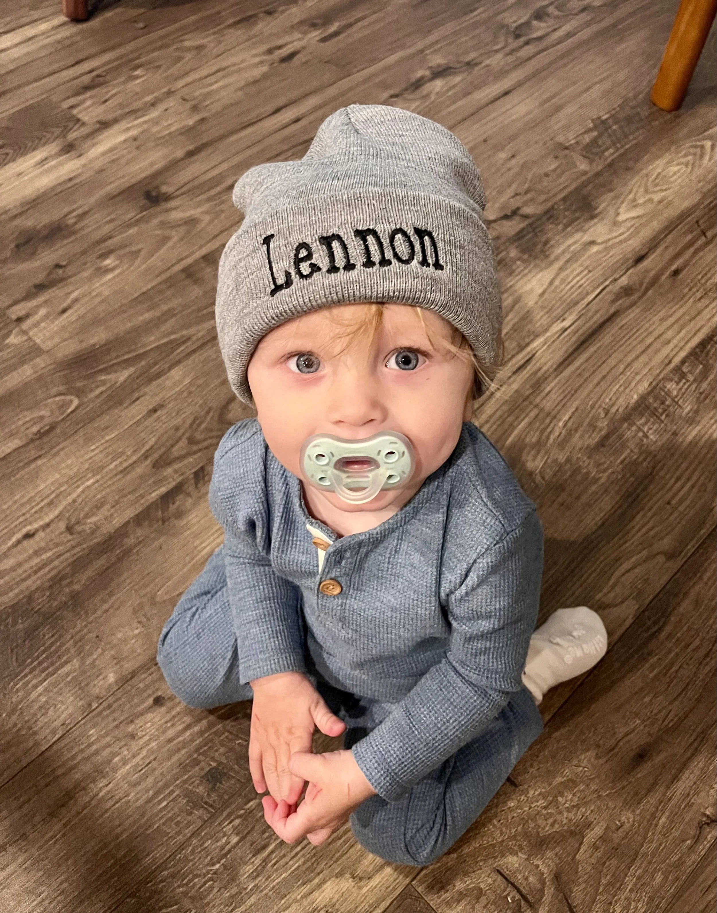 A young child with light brown hair and blue eyes, wearing a gray knit beanie with the word 'Lennon' embroidered on it, a gray button-up jacket, and a pacifier in their mouth. The child is sitting on a wooden floor, looking up at the camera, with an adult's hand gently holding their hand.