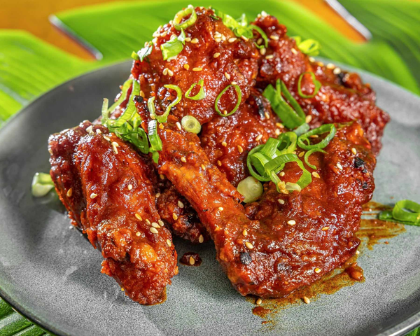 Fried chicken wings coated in spicy sauce, garnished with chopped green onions and sesame seeds on a black plate with green leaves in the background. Kaia's Kitchen Harristown