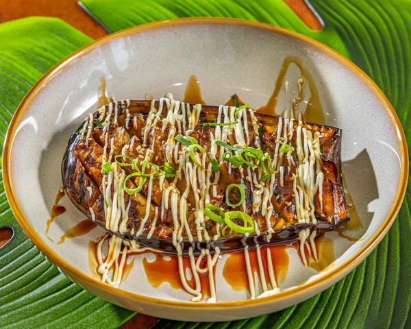 Grilled salmon fillet in a white bowl topped with white sauce, sliced green onions, and drizzled with soy sauce, sitting on a green tropical leaf background. Kaia's Kitchen Harristown