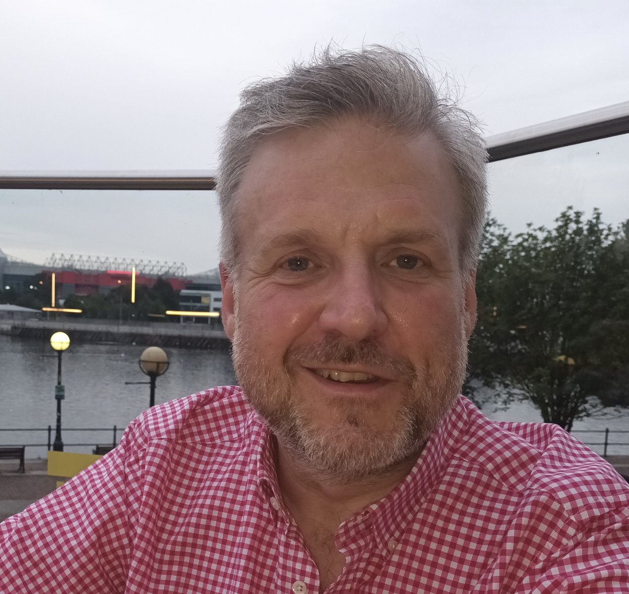 A smiling man with a beard and short gray hair wearing a red and white checkered shirt, outdoors near a body of water with trees and buildings in the background.