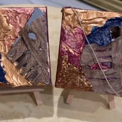Two abstract, textured mixed media artworks on small easels, featuring metallic and fabric elements.