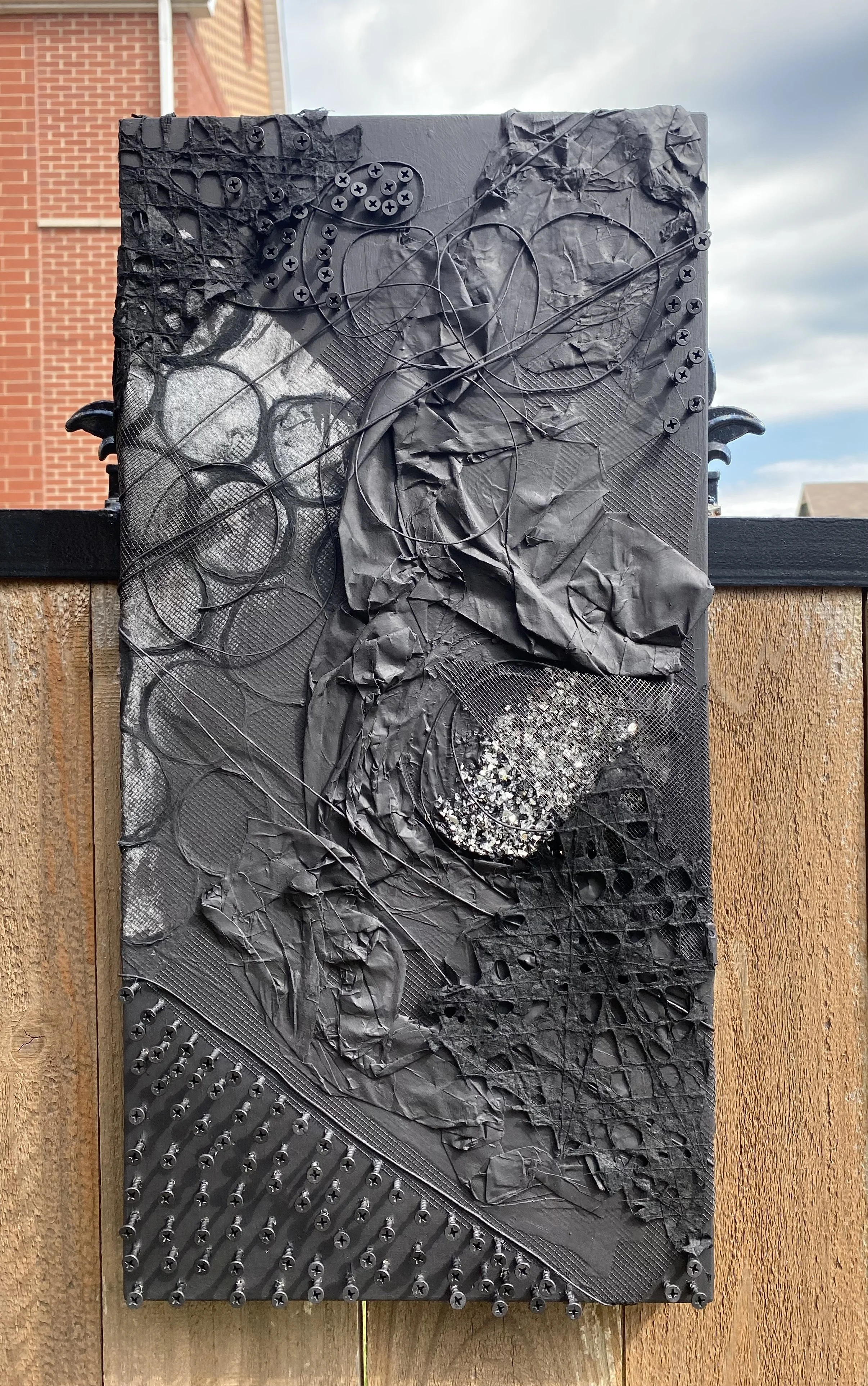 Black textured mixed media art piece with crumpled paper, wires, screws, and various materials arranged abstractly, mounted on a wooden outdoor fence with a cloudy sky and brick building in the background.