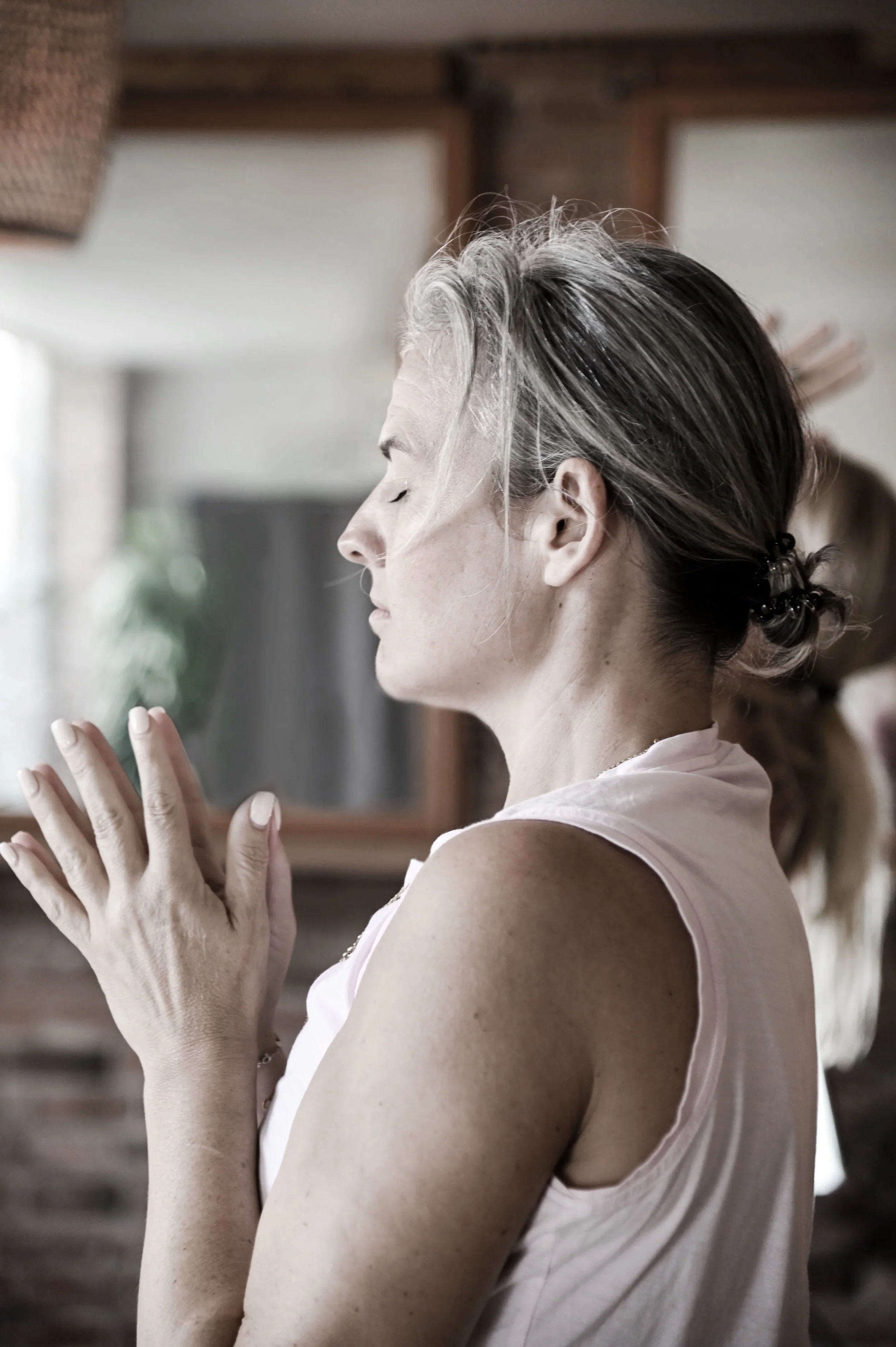 A woman with gray hair tied back in a bun, wearing a sleeveless top, has her eyes closed and hands pressed together in a prayer position indoors.