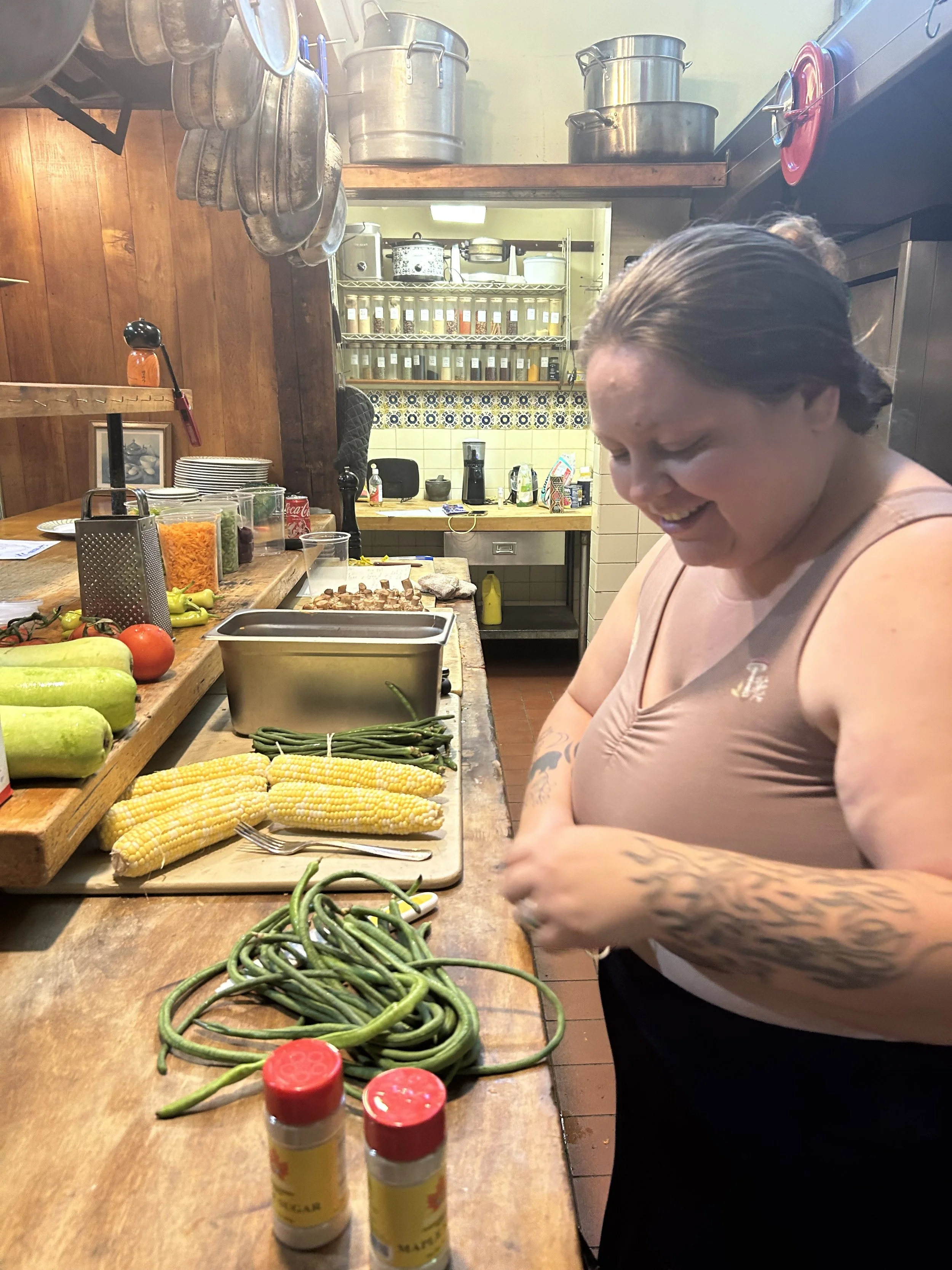 A woman in a kitchen with fresh vegetables including green beans, corn on the cob, and zucchini, preparing food with a smile.