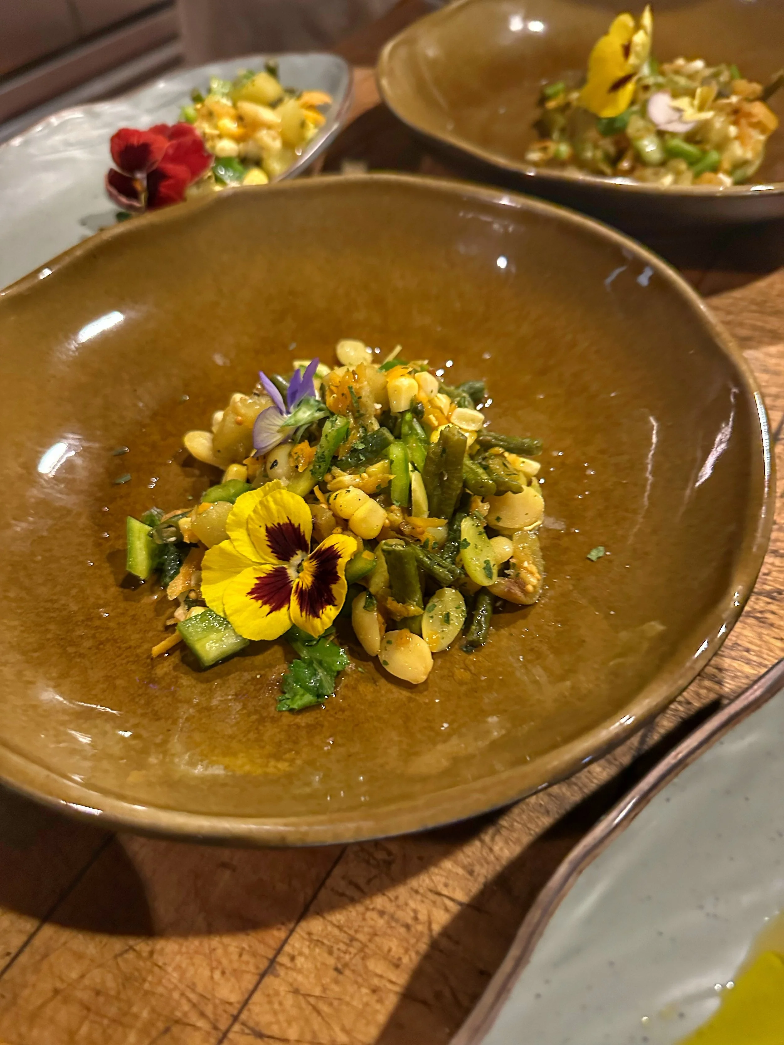 A dish of mixed vegetables garnished with edible flowers, served in a brown ceramic bowl.