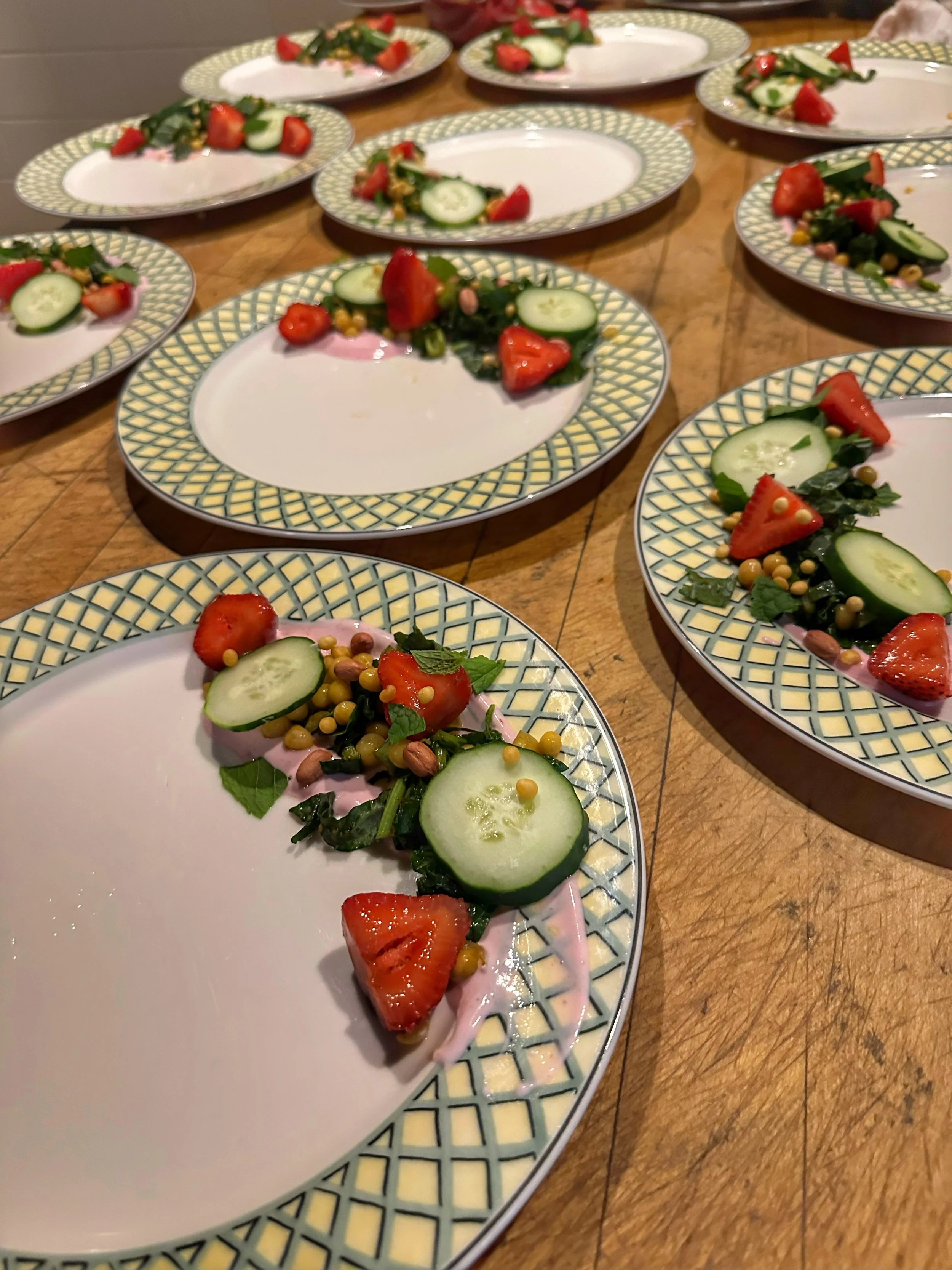 Empty plates with salad containing strawberries, cucumber slices, leafy greens, and small yellow, pink, and purple seeds or pulses, arranged on a wooden table.