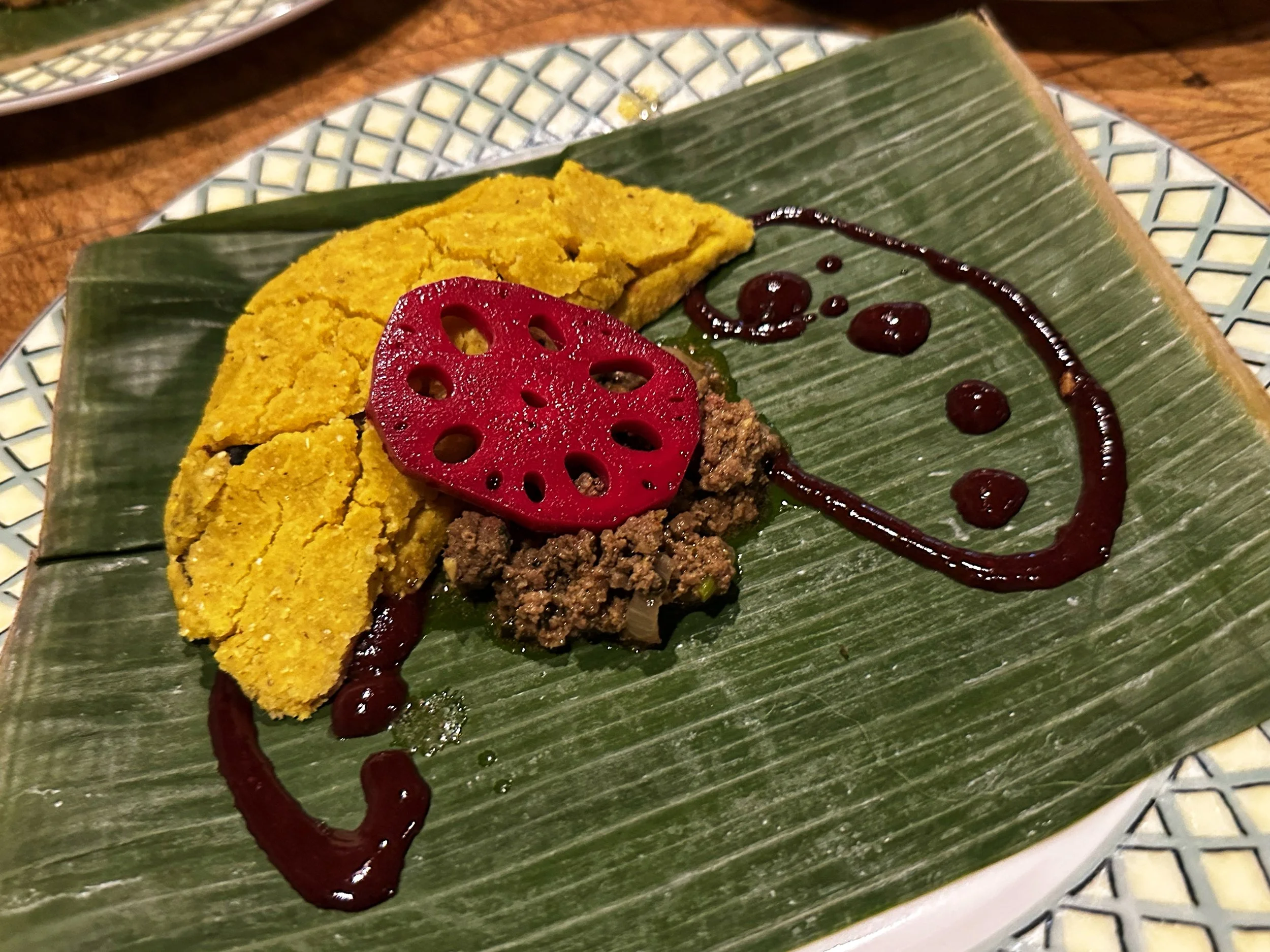 A dessert on a green leaf placed on a patterned plate, consisting of yellow cake, a sliced red lotus root, ground nuts, and a drizzle of chocolate sauce arranged in a smiley face shape.