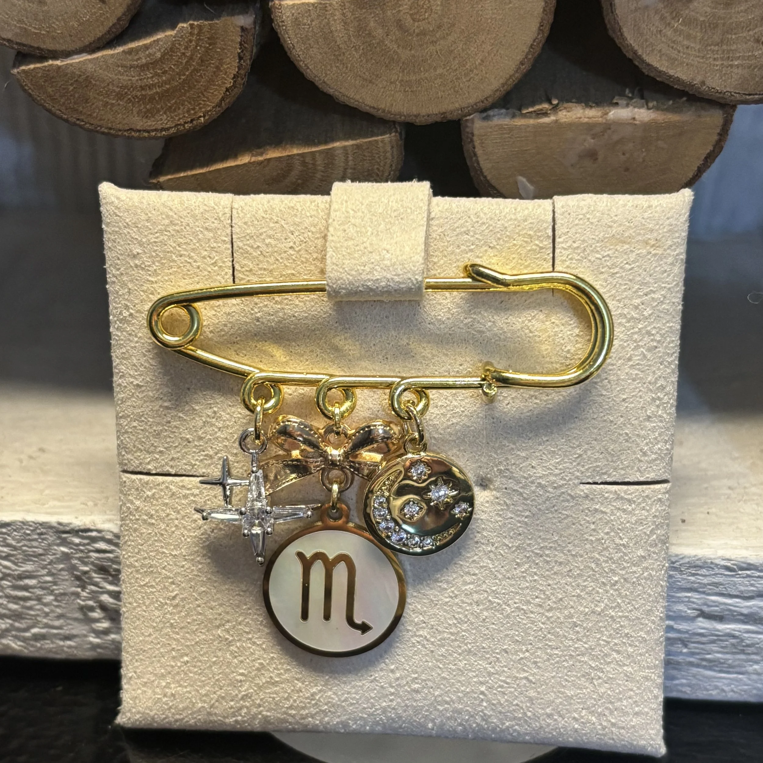 Broche de taille moyenne en forme de épingle dorée avec cinq breloques suspendues : une étoile en argent, une fleur avec des cristaux, une lune avec des cristaux et une étoile, une pièce ronde avec le signe astrologique du Scorpion, et une demi-lune avec un visage de lune et des cristaux. Tous accrochés à la broche.