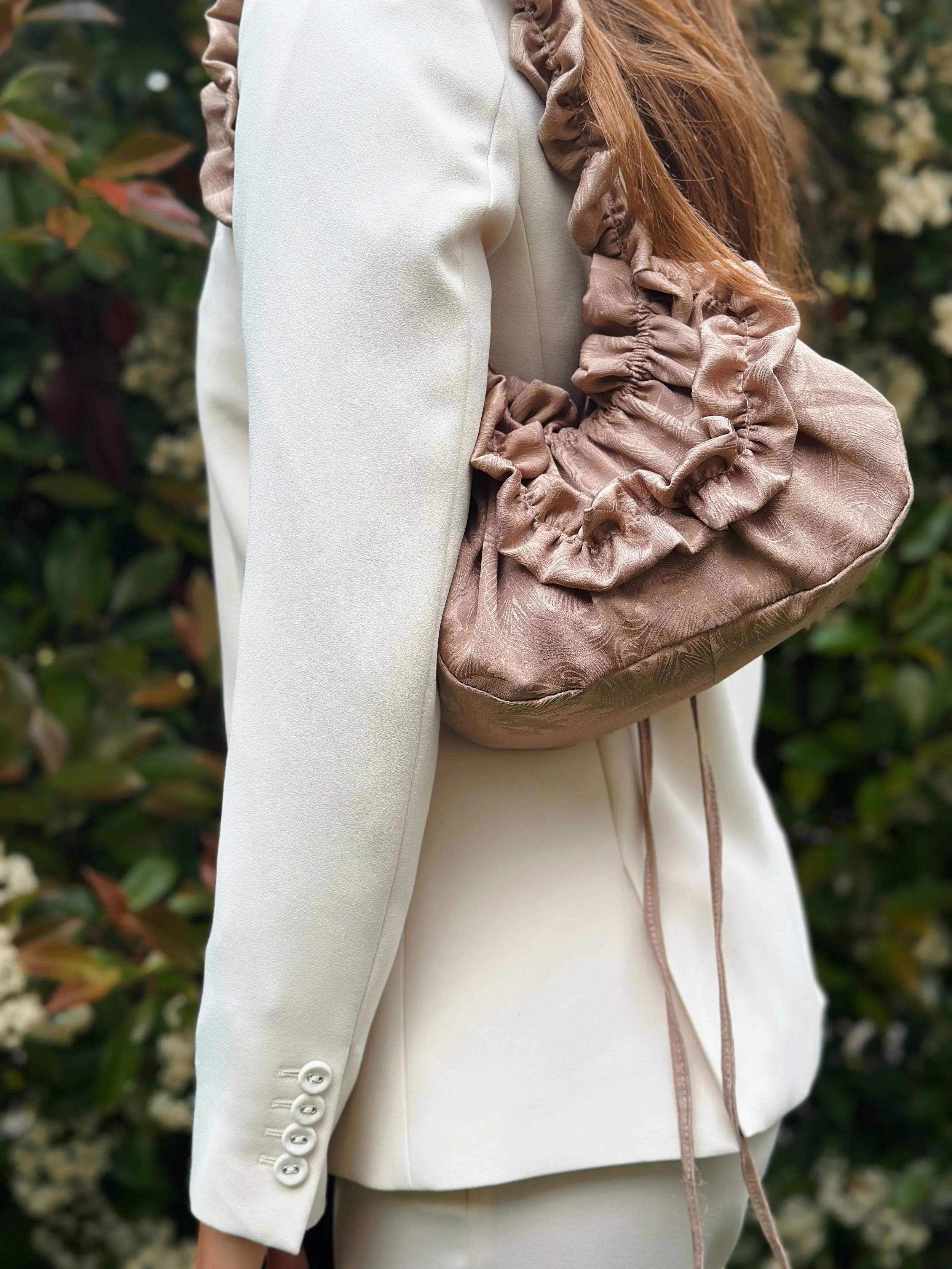 Une personne porte une veste blanche avec des boutons aux manches, et tient un sac à main en tissu beige avec des plissures et des rubans attachés, dans un environnement végétal.