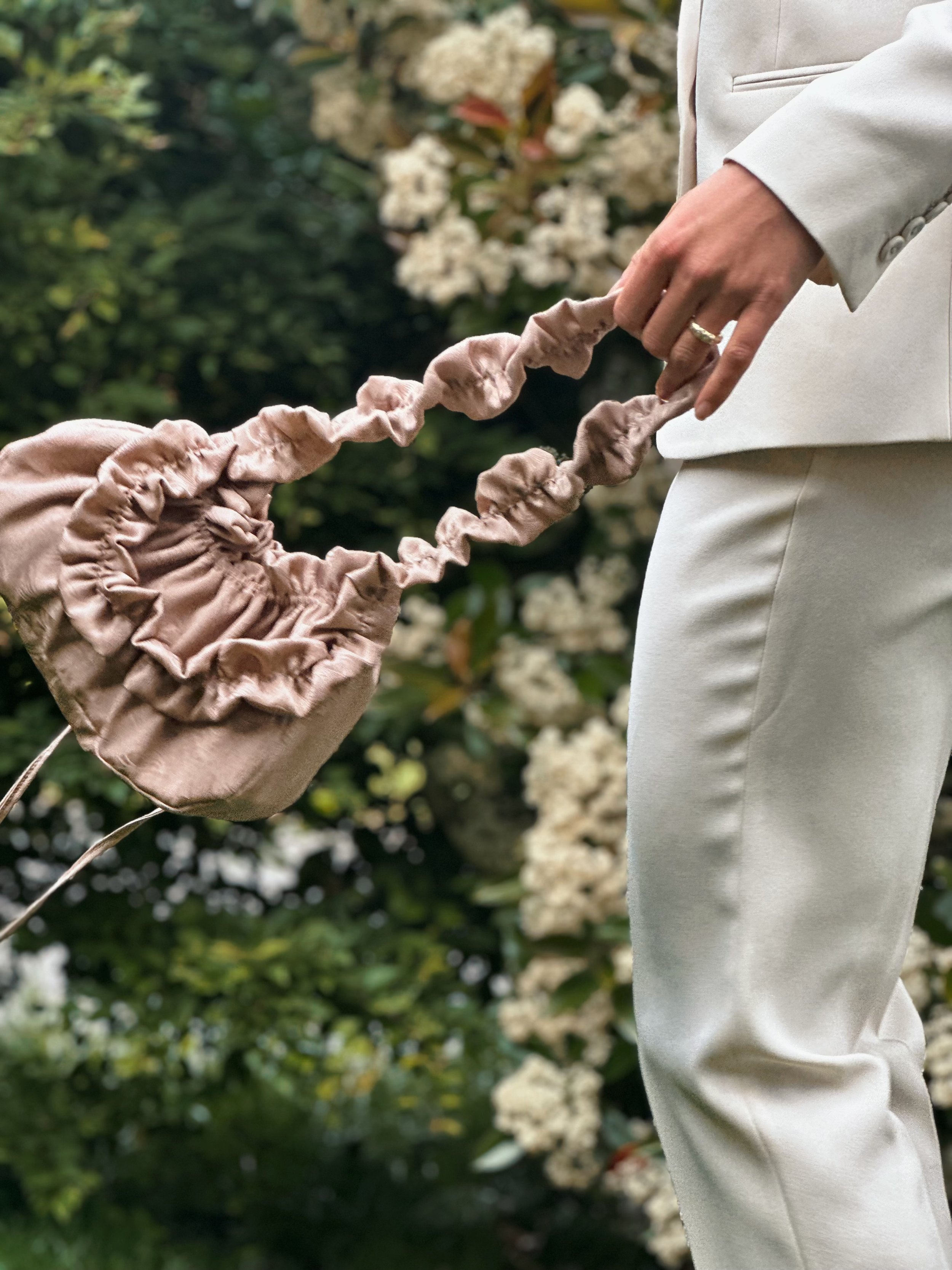 Une personne habillée en blanc tient un sac en tissu beige avec une bandoulière ruffled, dans un jardin fleuri.