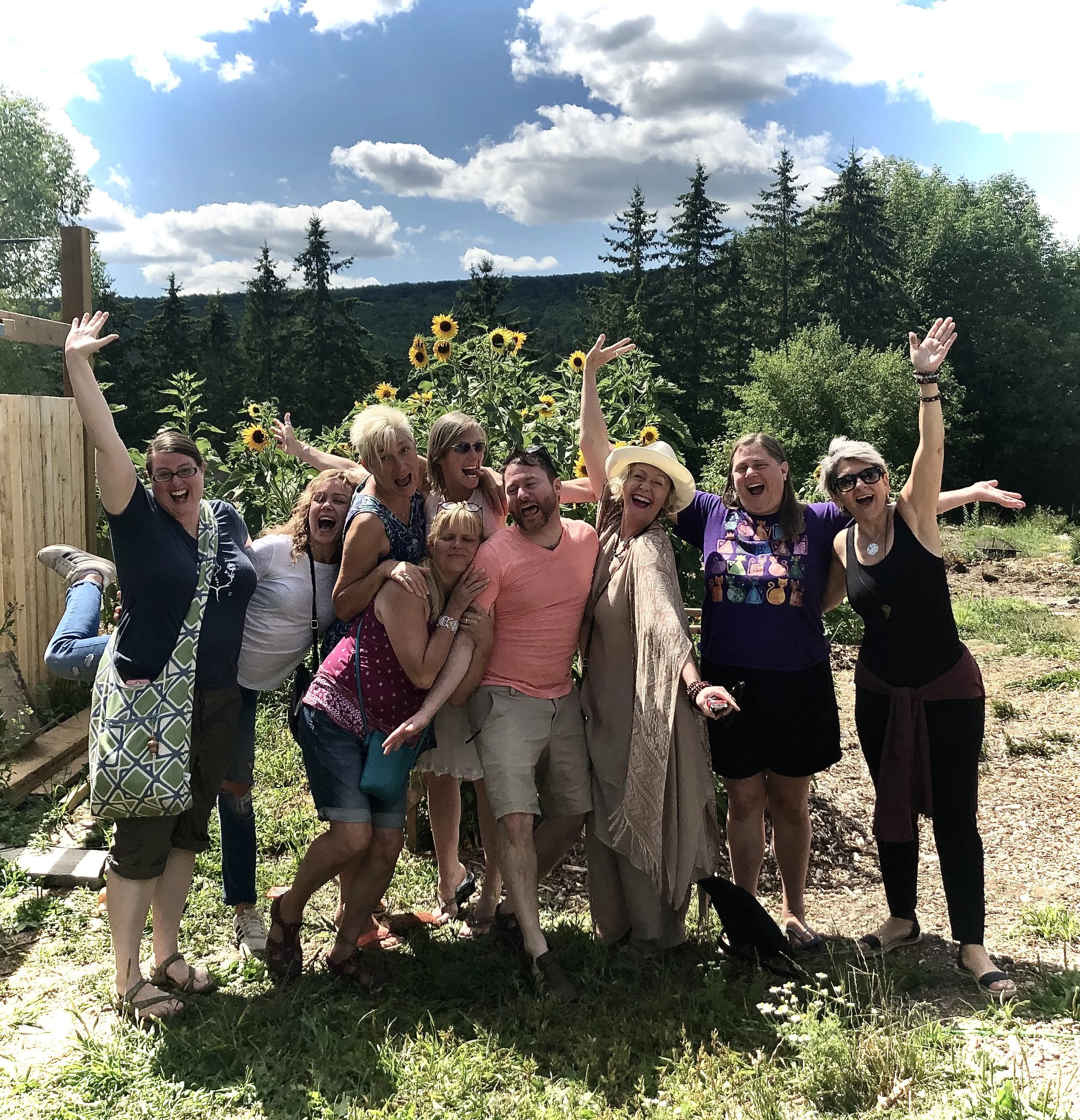 Group of smiling people standing outdoors with AdiKanda, near sunflower plants, posing cheerfully with arms raised and arms around each other.