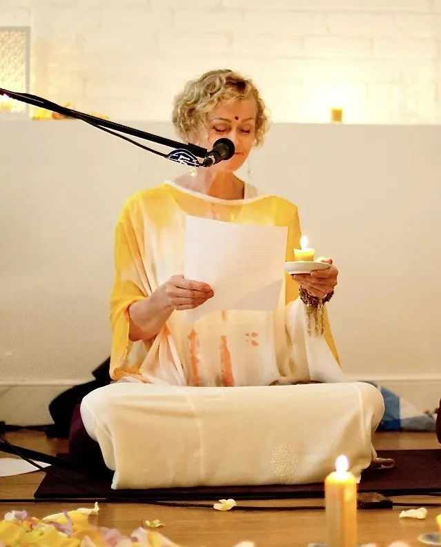 AdiKanda sitting cross-legged on the floor, reading a transmission from a piece of paper, with glowing candles and flower petals around her, in a peaceful indoor setting.