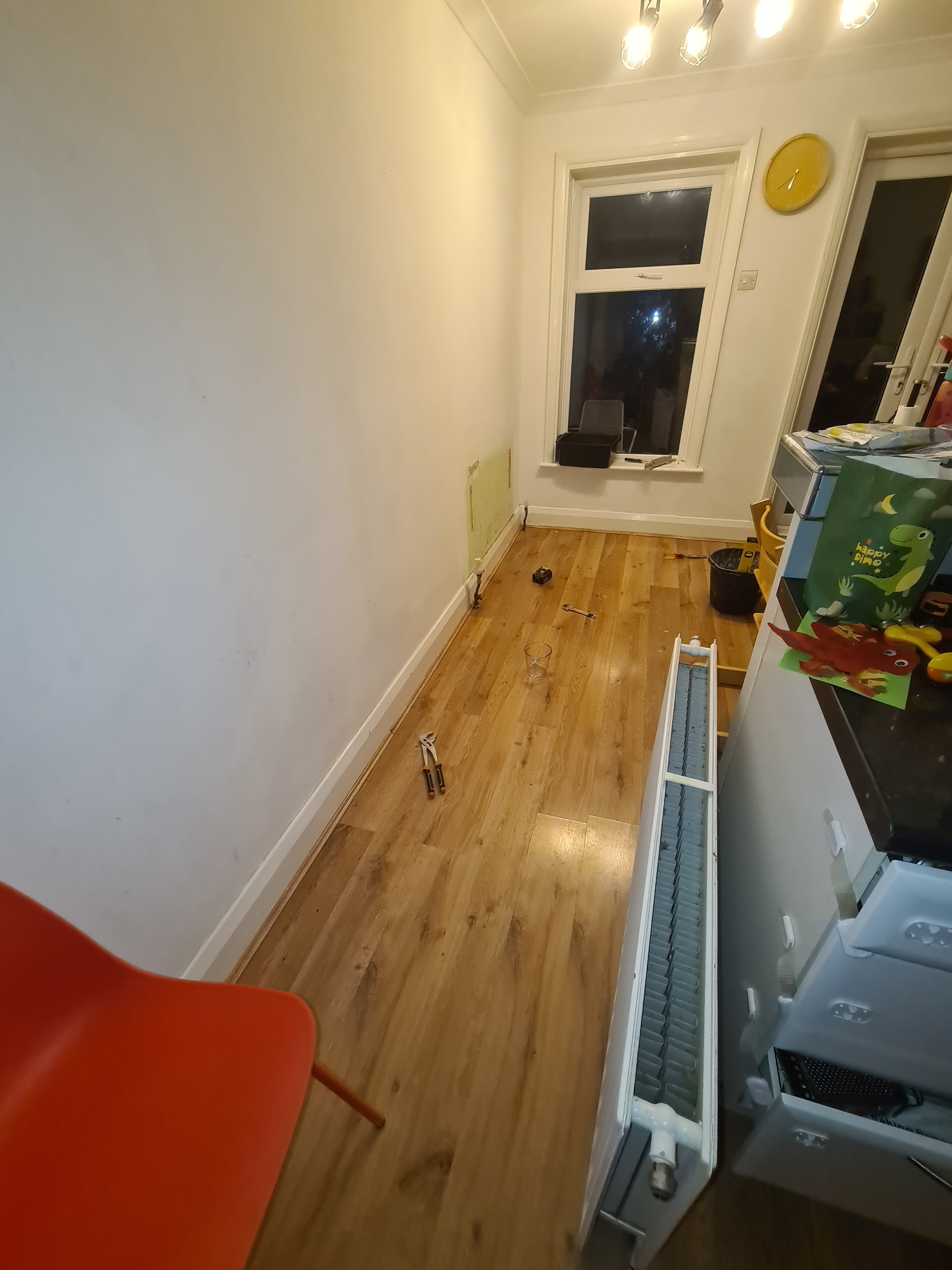A narrow kitchen with wooden flooring, a white radiator on the right, a window with a black object on the sill, a yellow wall clock, and some scattered tools and objects on the floor. There's an orange chair in the corner and various items on the bla