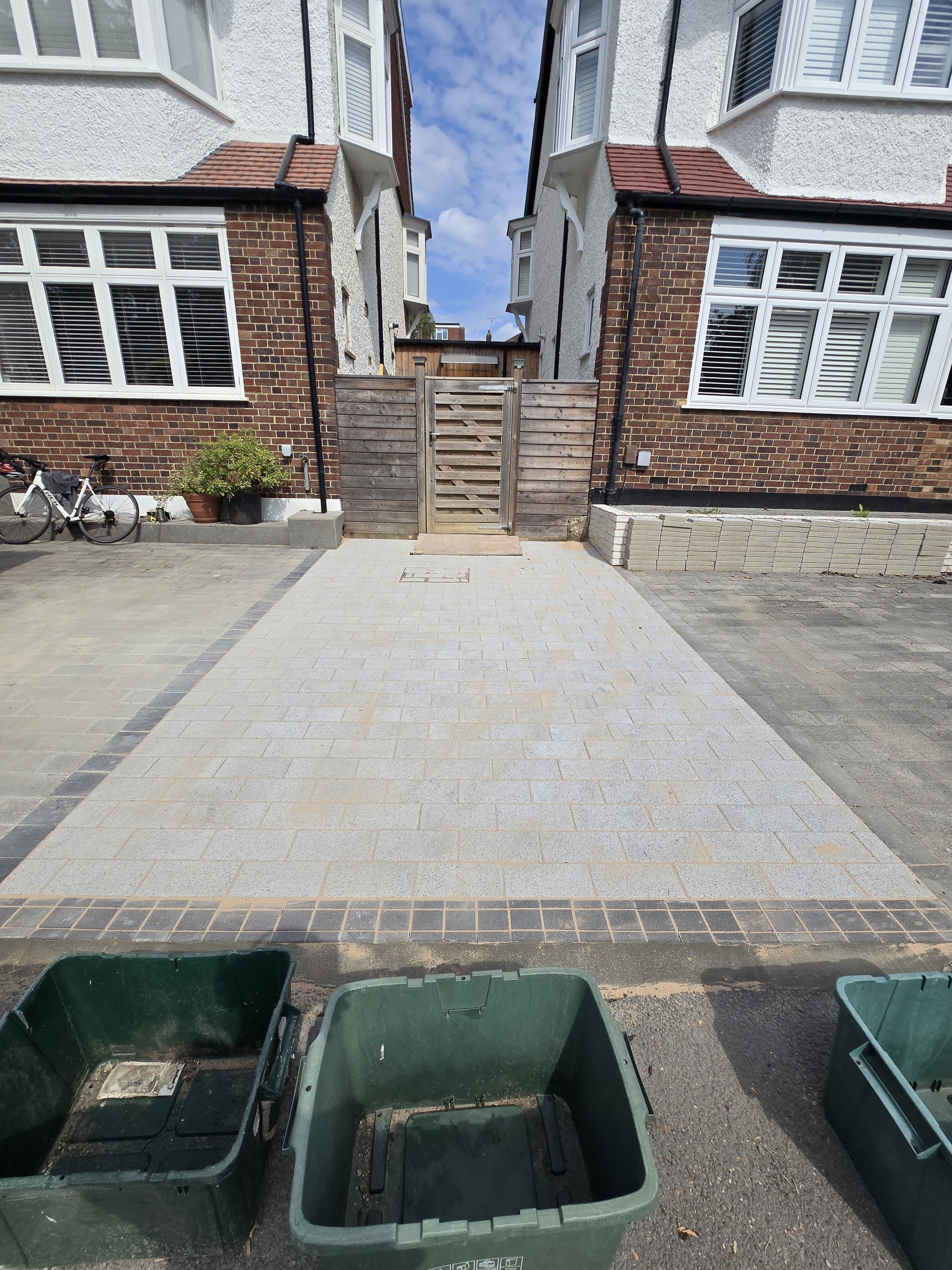 A view of a residential driveway with light gray paving stones, bordered by darker bricks. Two green waste bins are at the front, and there is a wooden gate at the back. The house has large windows with white frames, brick and white stucco exterior, 