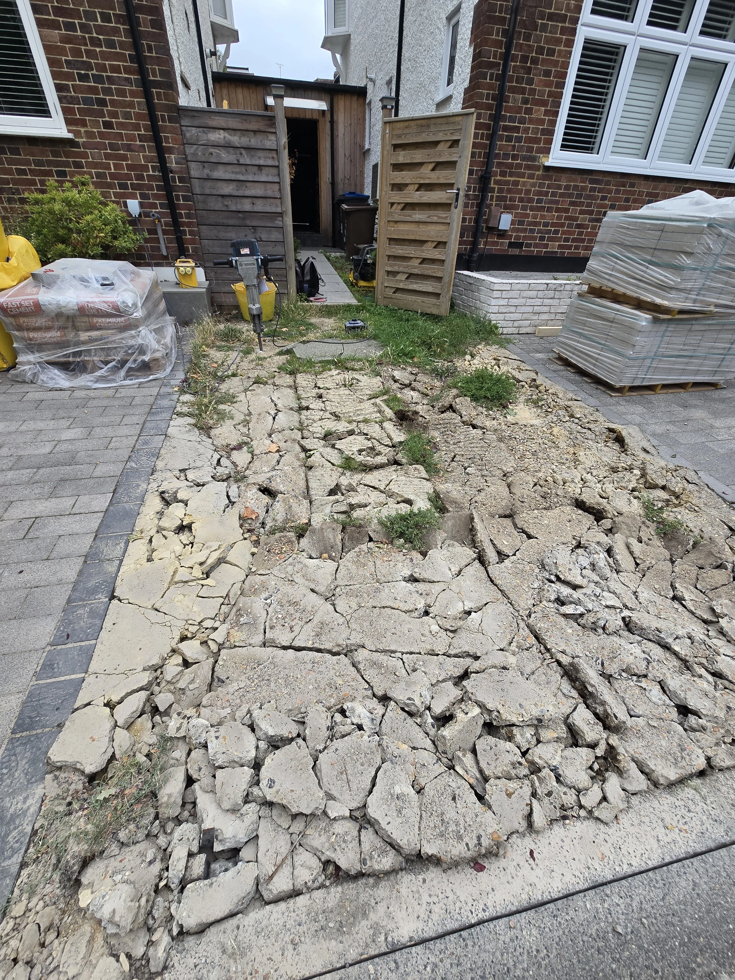 A residential backyard with a section of cracked, broken concrete pavement being removed. Piles of new paving stones are stacked on the side, and construction tools are present. The yard is enclosed by a wooden gate and fencing, with garden and build