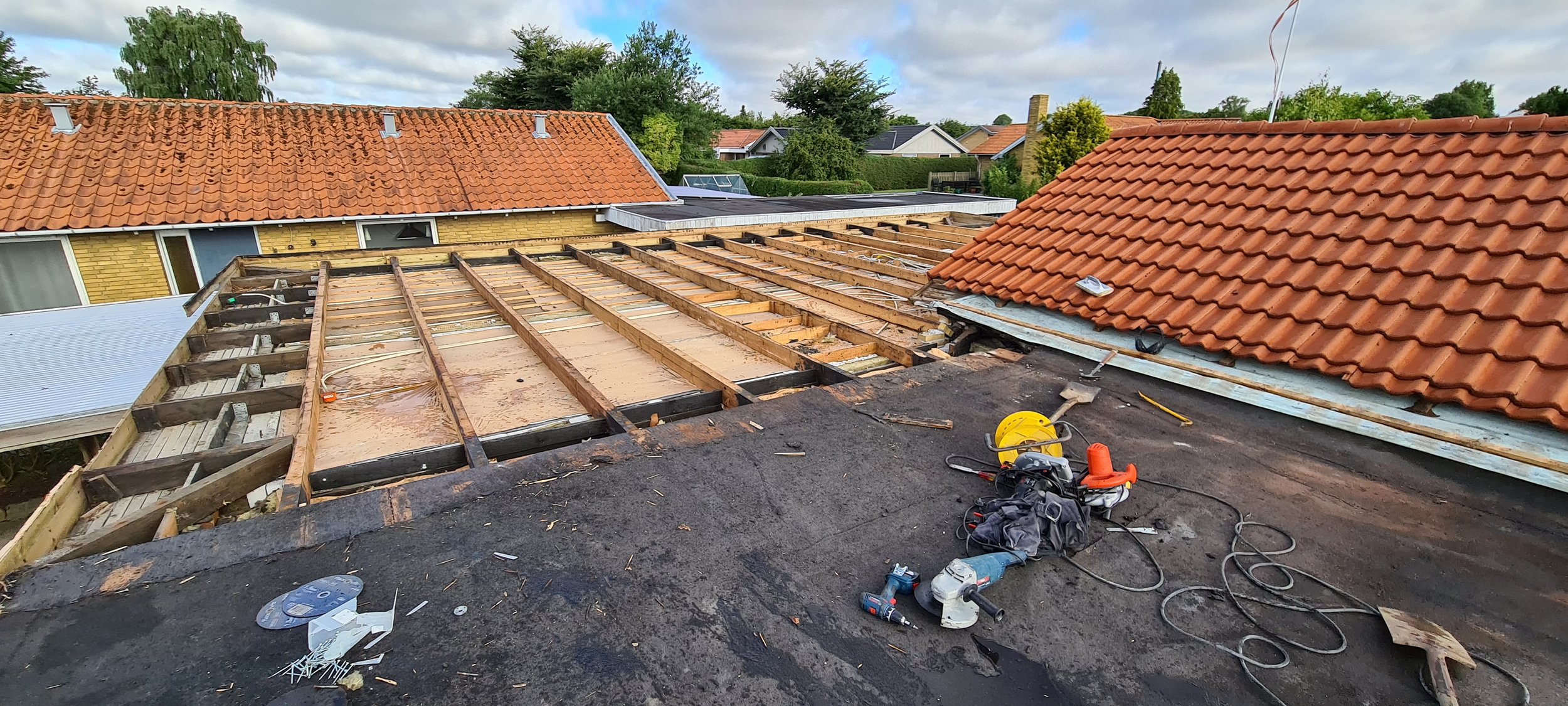 Roof under construction with exposed wooden framework, construction tools including a drill, circular saw, extension cord, and various other supplies scattered on the black roofing material. Adjacent roof with red tiles is mostly completed.