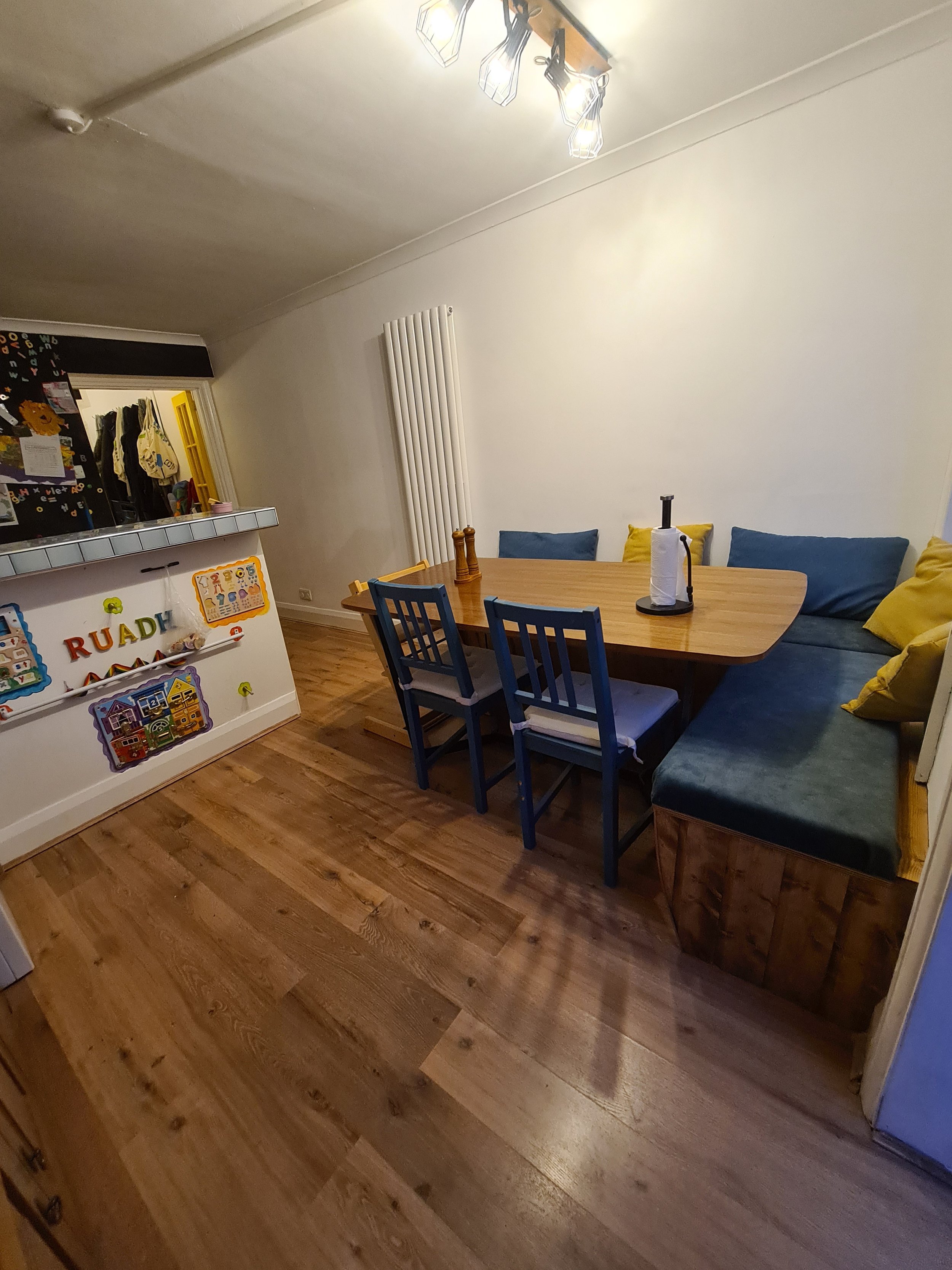 A dining room with a wooden table, blue and yellow pillows on a built-in corner bench, and six chairs, three on each side. There is a paper towel holder and a bottle on the table. The floor is wooden, and there is a white radiator on the wall. Overhe
