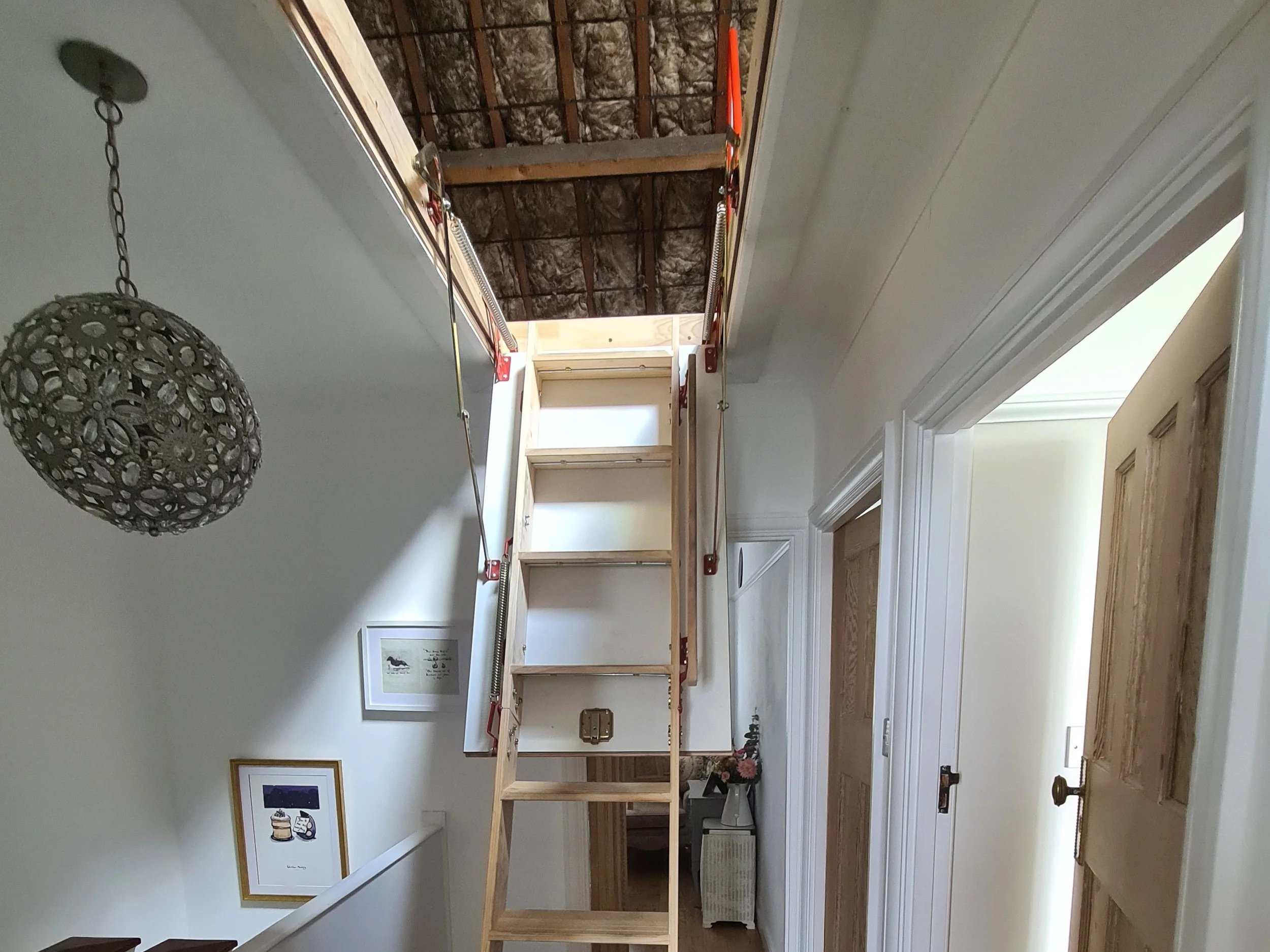 Interior view of a house under renovation showing a staircase with wooden steps and a ladder leading up to an unfinished attic space with exposed insulation and beams.