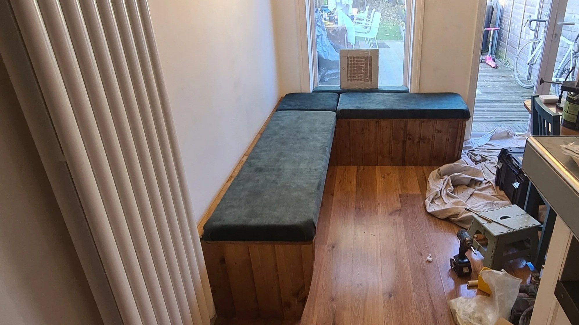 A corner built-in bench with green upholstery, wooden base, and a cushion on top, situated next to a sliding glass door leading to an outdoor patio. The patio has plastic chairs, a table, and bicycles, with tools and cleaning supplies scattered on th