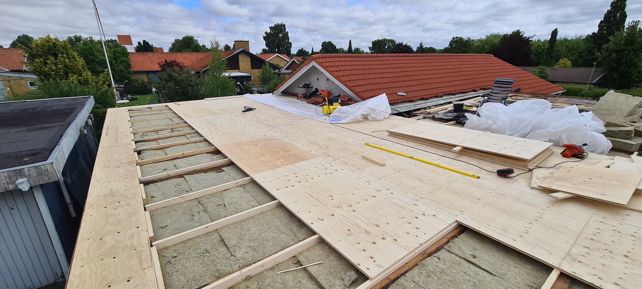 Rooftop under construction with plywood sheets and tools laid out, neighboring houses with trees and cloudy sky in the background.