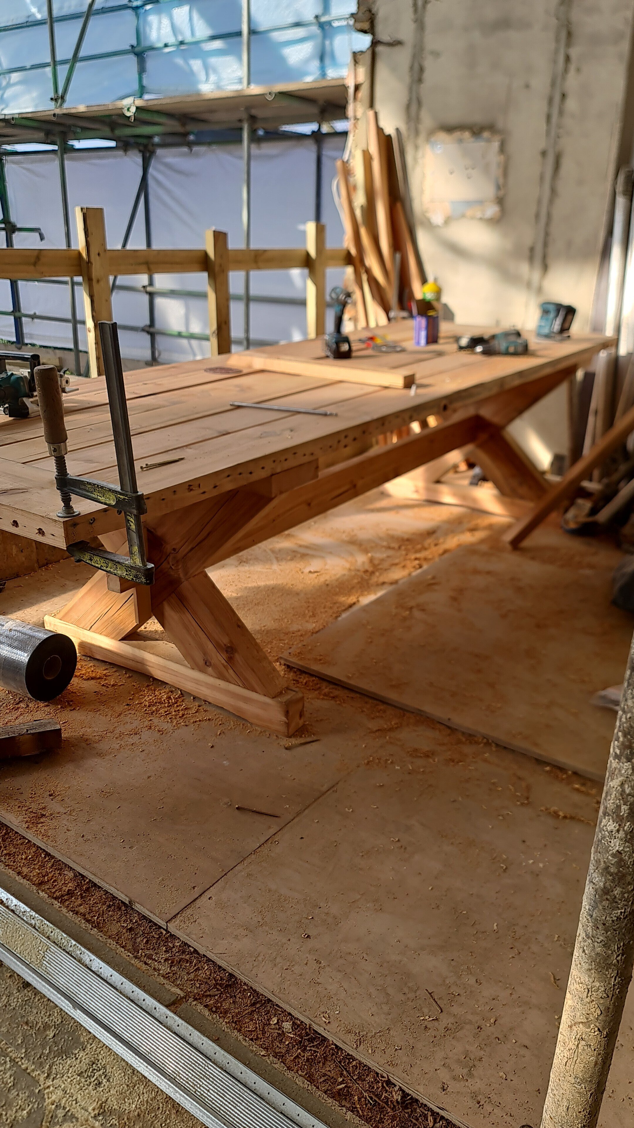 Wooden workbench in an unfinished construction site, with sawdust, tools, and wood pieces on it, surrounded by scaffolding and a partially completed wall.