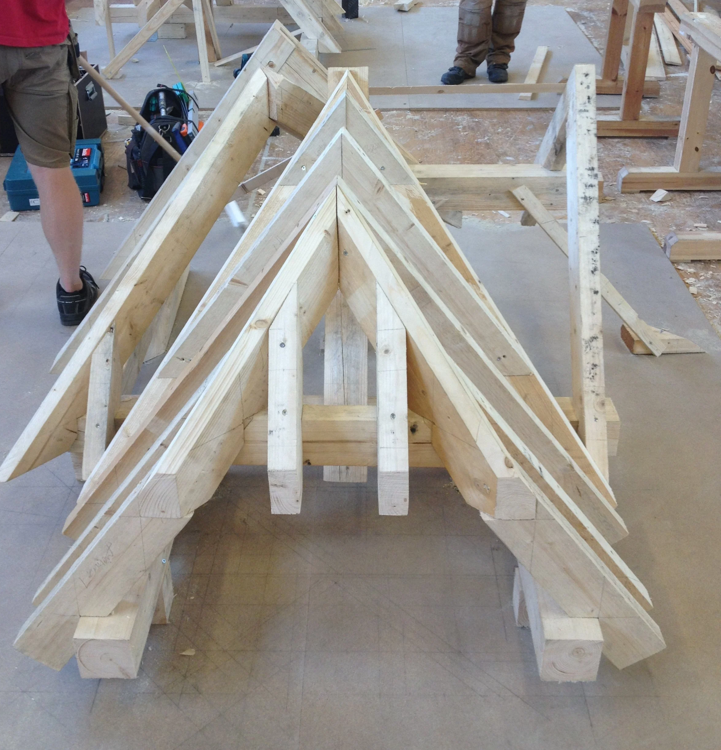 Wooden framing for a roof being constructed with multiple triangular trusses made of light-colored wood, in a workshop setting.